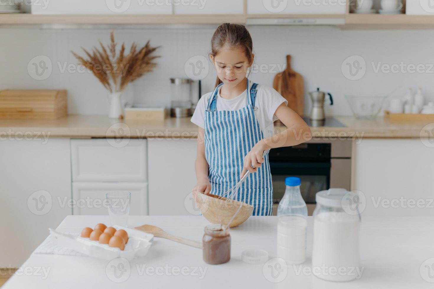 Small cute girl in apron, mixes ingredients, whisks with beater, uses eggs, milk, flour, tries new recipe, stands against kitchen interior, prepares tasty cookies or bakery, learns how to cook. photo