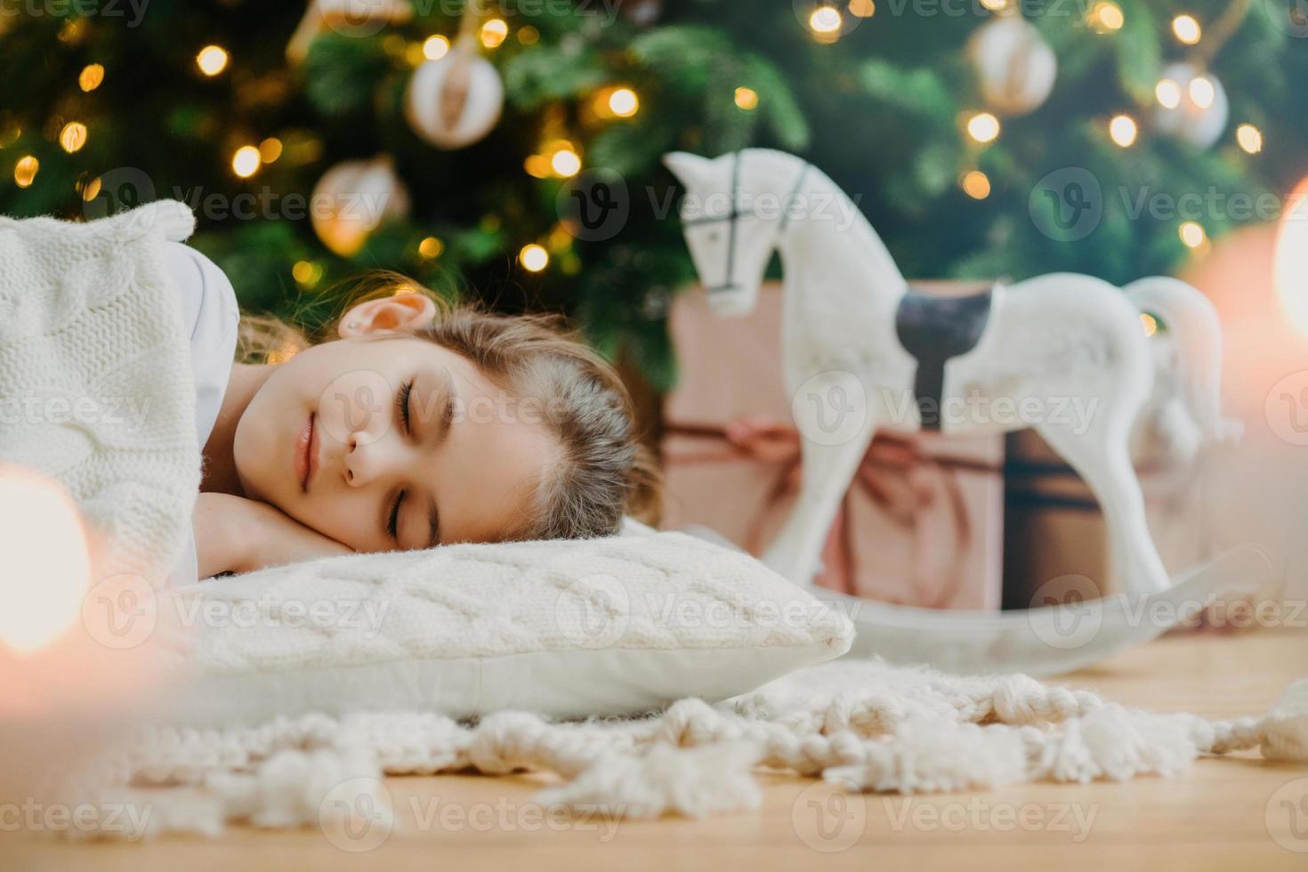 Horizontal shot of relaxed girl covered with white sweater, has pleasant dreams, lies under decorated Christmas tree, toy rocker and gift boxes around. Kid at home. Cozy holiday. Domestic atmosphere photo