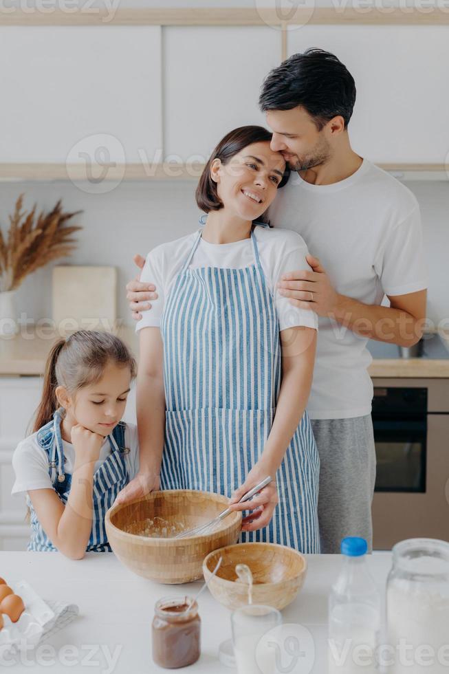 familia cocina comida en casa. el esposo y la esposa se abrazan con amor, la niña pequeña posa cerca, prepara una cena deliciosa, usa leche, chocolate, huevos, hace pasteles caseros, posa contra el interior de la cocina. foto