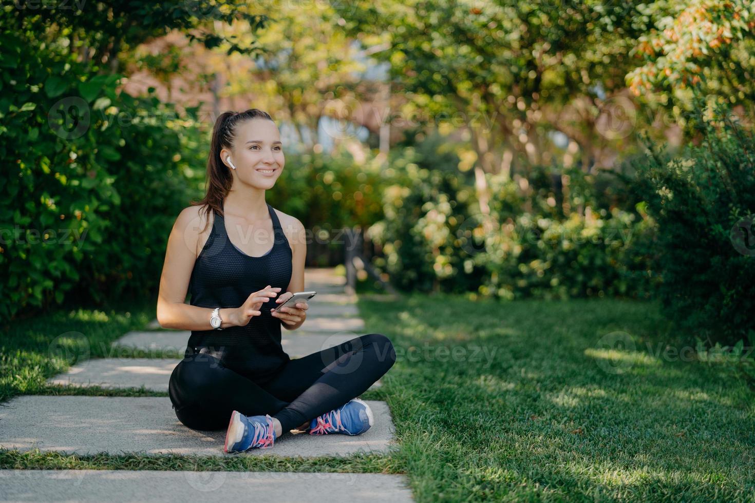 Cheerful female runner poses in lotus pose outdoor dressed in sportswear uses mobile phone during rest after jog downloads music songs in player prepares for morning workout chooses audio records photo