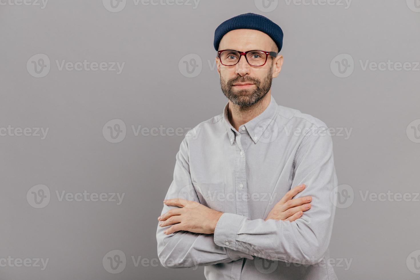 Photo of serious male keeps arms folded over chest, dressed in white formal shirt, fashionable hat, poses for making photography over grey wall with free space for your advertisement or promotion