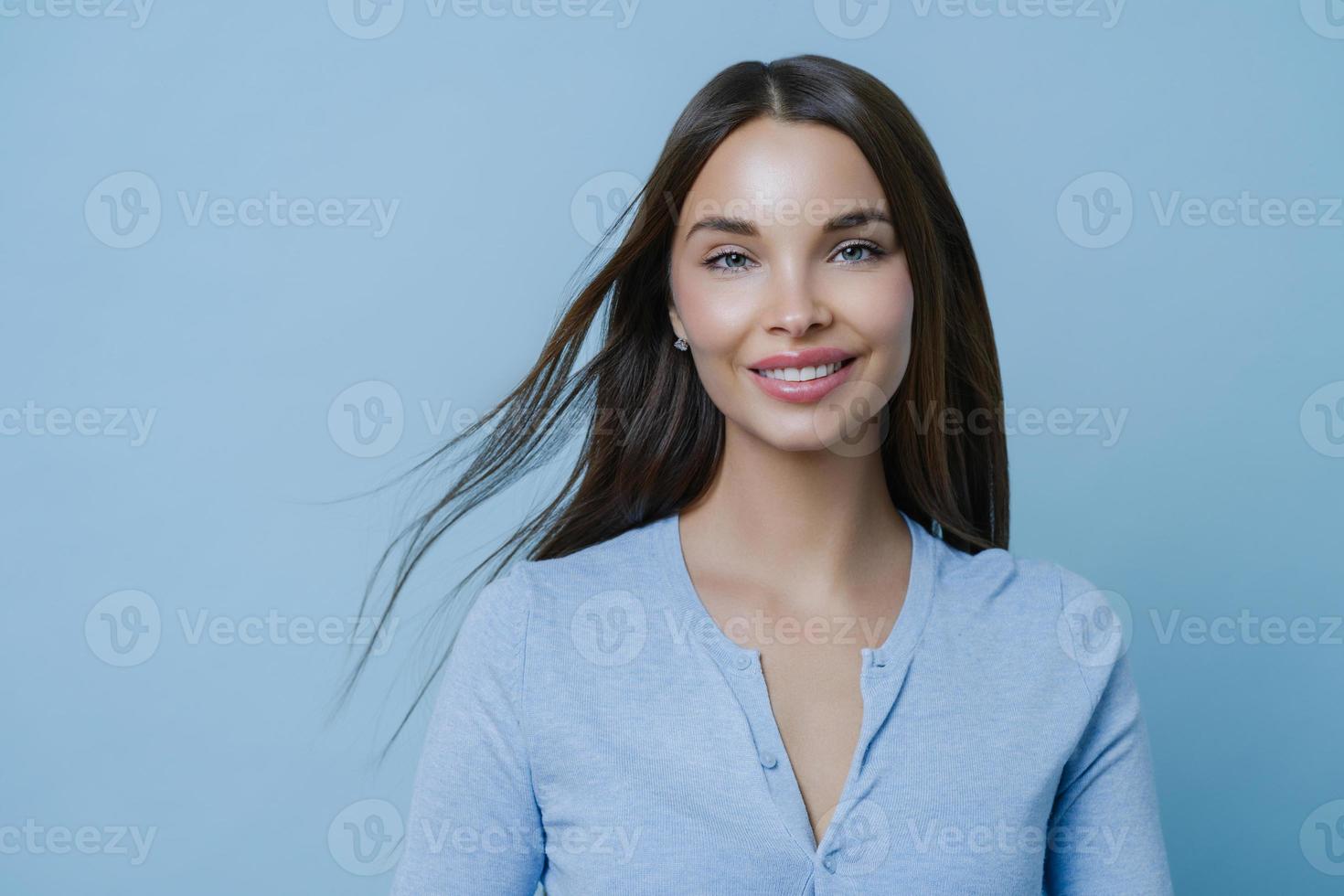 Positive lovely feminine lady happy to have first date with boyfriend, smiles broadly, has appealing appearance, glad to hear something pleasant, models indoor alone, wears blue casual jumper photo