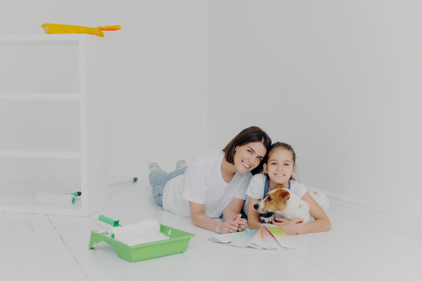 Joyful friendly family members with dog lie on floor in empty room which needs improvement, choose color on palette sample, finish house redecoration, have good relationship. Teamwork, house chores photo