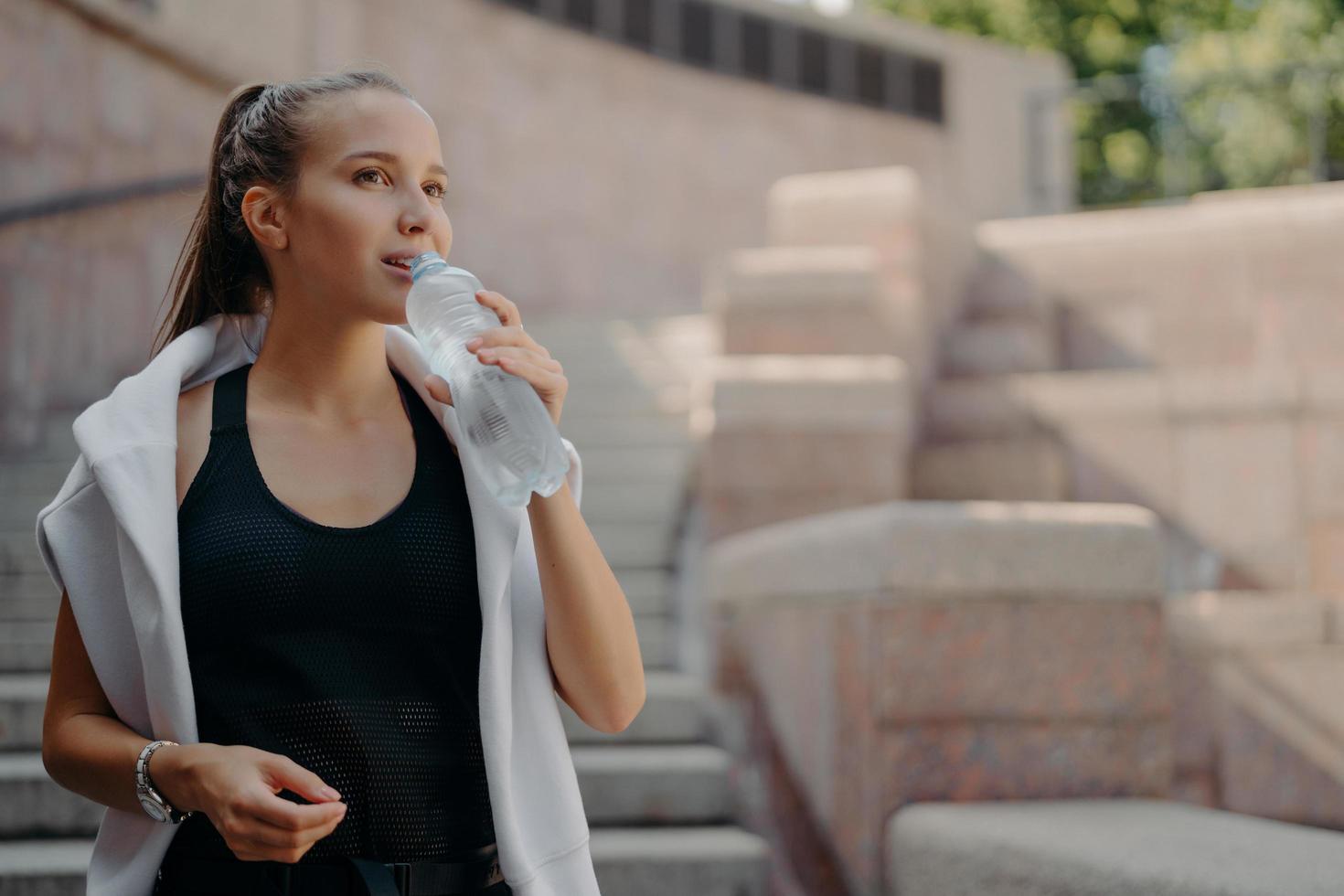 mantener el equilibrio del agua. una joven deportista exhausta bebe una bebida fría y refrescante después de que el entrenamiento toma un descanso, usa una camiseta y un suéter blanco alrededor de los hombros, siente sed. concepto de deporte y salud foto