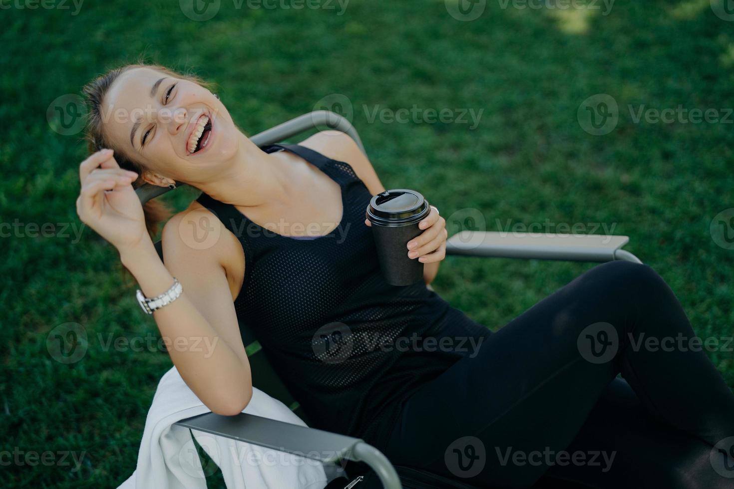 Overjoyed relaxed woman smiles broadly enjoys drinking coffee outdoor dressed in t shirt and trousers poses in comfortable chair against green grass. View from above. People leisure lifestyle photo