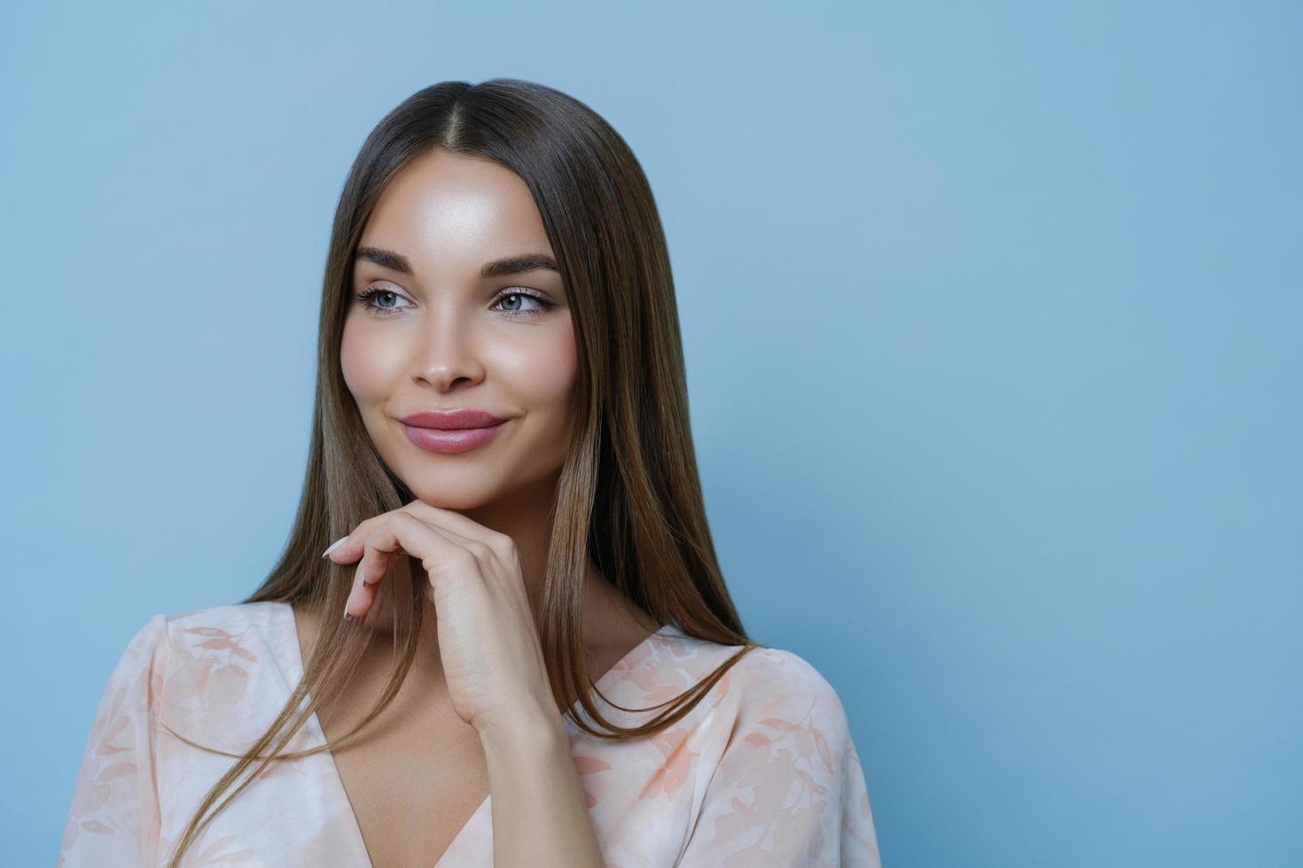 Portrait of good looking woman touches chin and has thoughtful expression, looks away, isolated over blue background, blank space for your advertisement or promotion. Romance and beauty concept photo