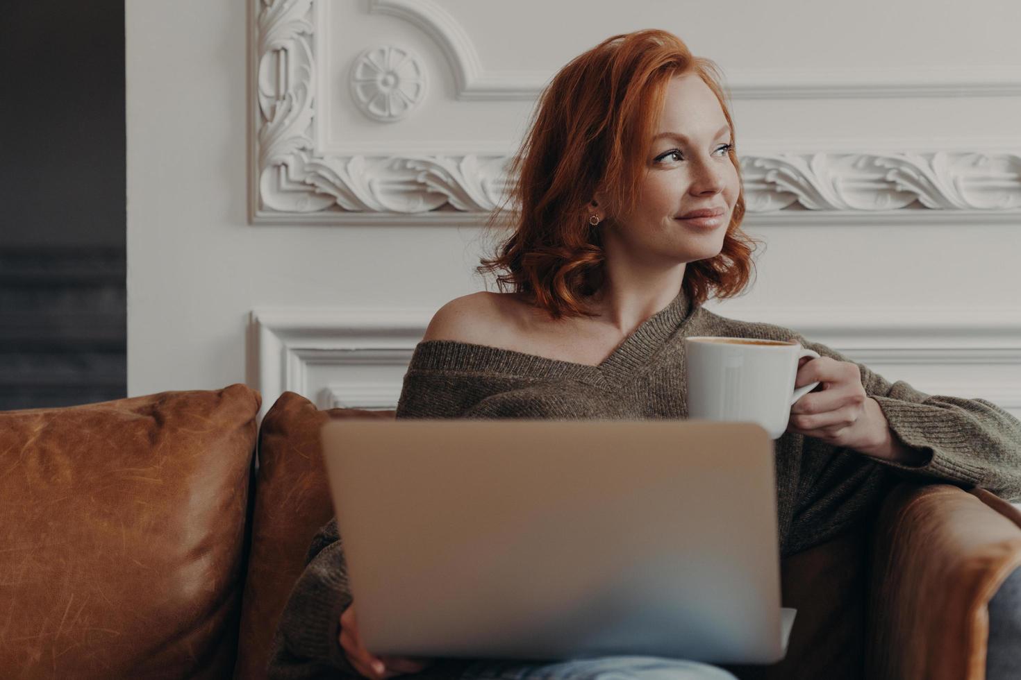 Indoor shot of satisfied redhead woman works freelance on laptop computer, drinks aromatic beverage, concentrated aside with thoughtful expression, sits on comfortable sofa, thinks about new project photo