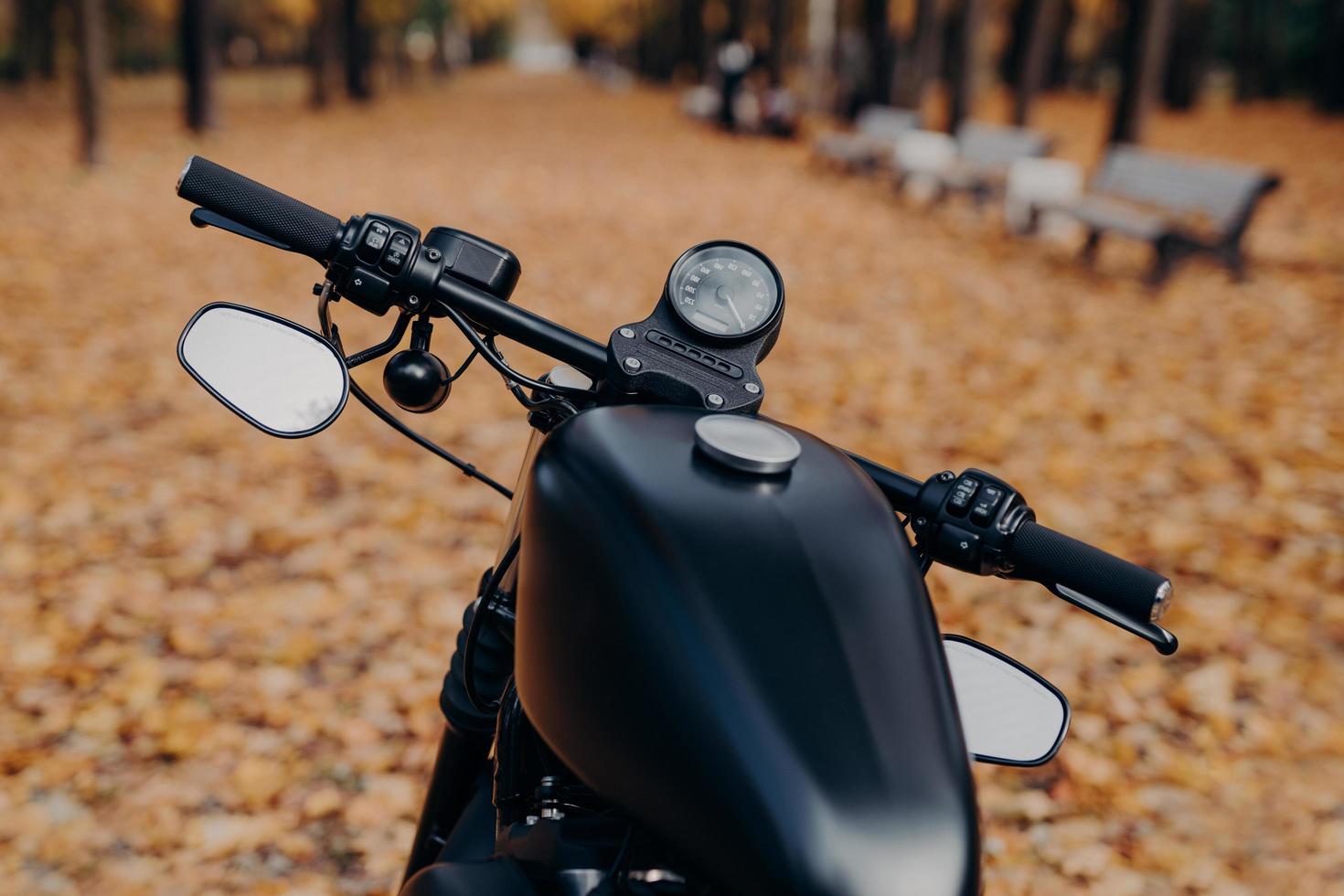 Close up shot of black motorcycle with speedometer, handlebar stands in autumn park against orange fallen leaves and benches. Transport concept. Bike parked outdoor photo
