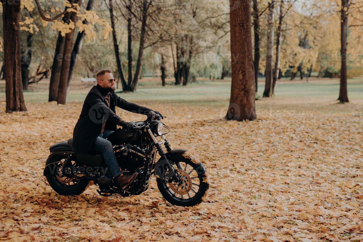 Confident male biker sits on motorbike, enjoys traveling on own transport, wears protective sunglasses, comfortable clothes, rides through trees in autumn park. Fall season, hobby, lifestyle photo