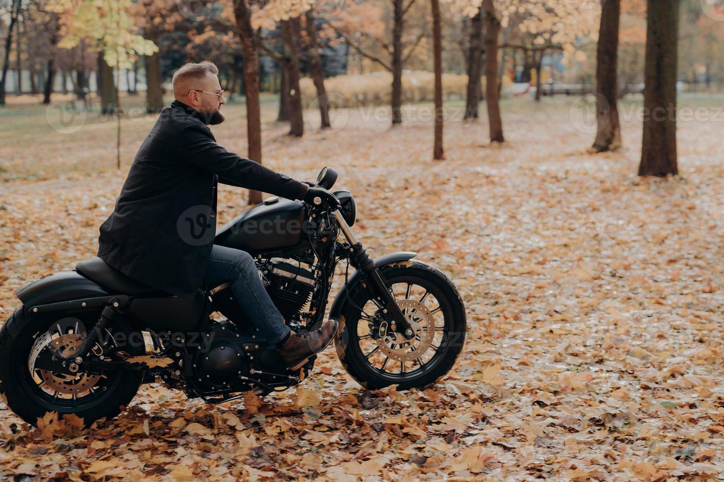 Horizontal shot of bearded biker in action or movement against park during autumn time, going to reach destination quickly, wears sunglasses, jacket, jeans and shoes, has adventurous trip alone photo