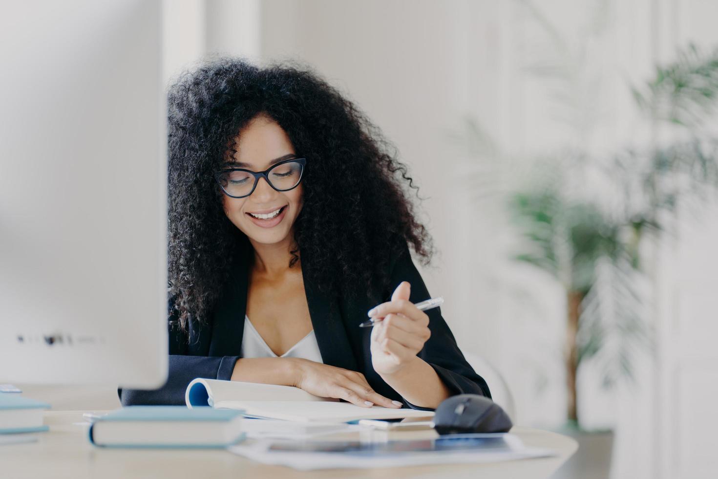 Photo of pleased curly haired woman writes down some information, holds pen, has smile, wears optical glasses and formal clothes poses at workplace with computer. Student writes ideas for course paper