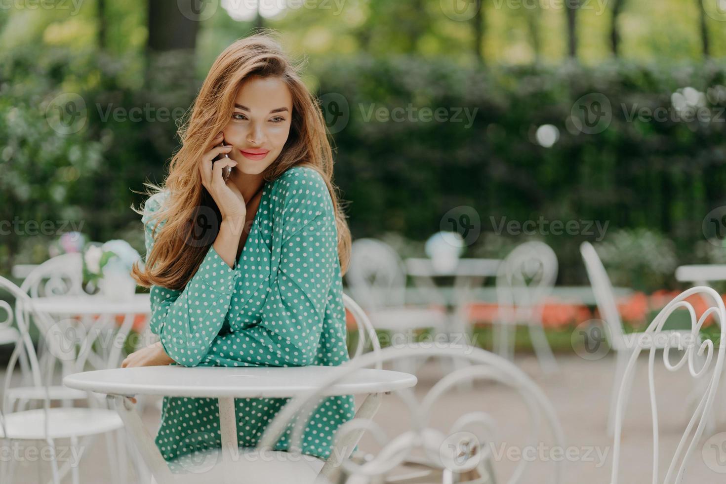 Outside shot of charming young woman with long hair, wears polka dot green shirt, sits at table in outdoor cafe, has pleasant talk via modern smartphone, has dreamy expreession. People and lifestyle photo