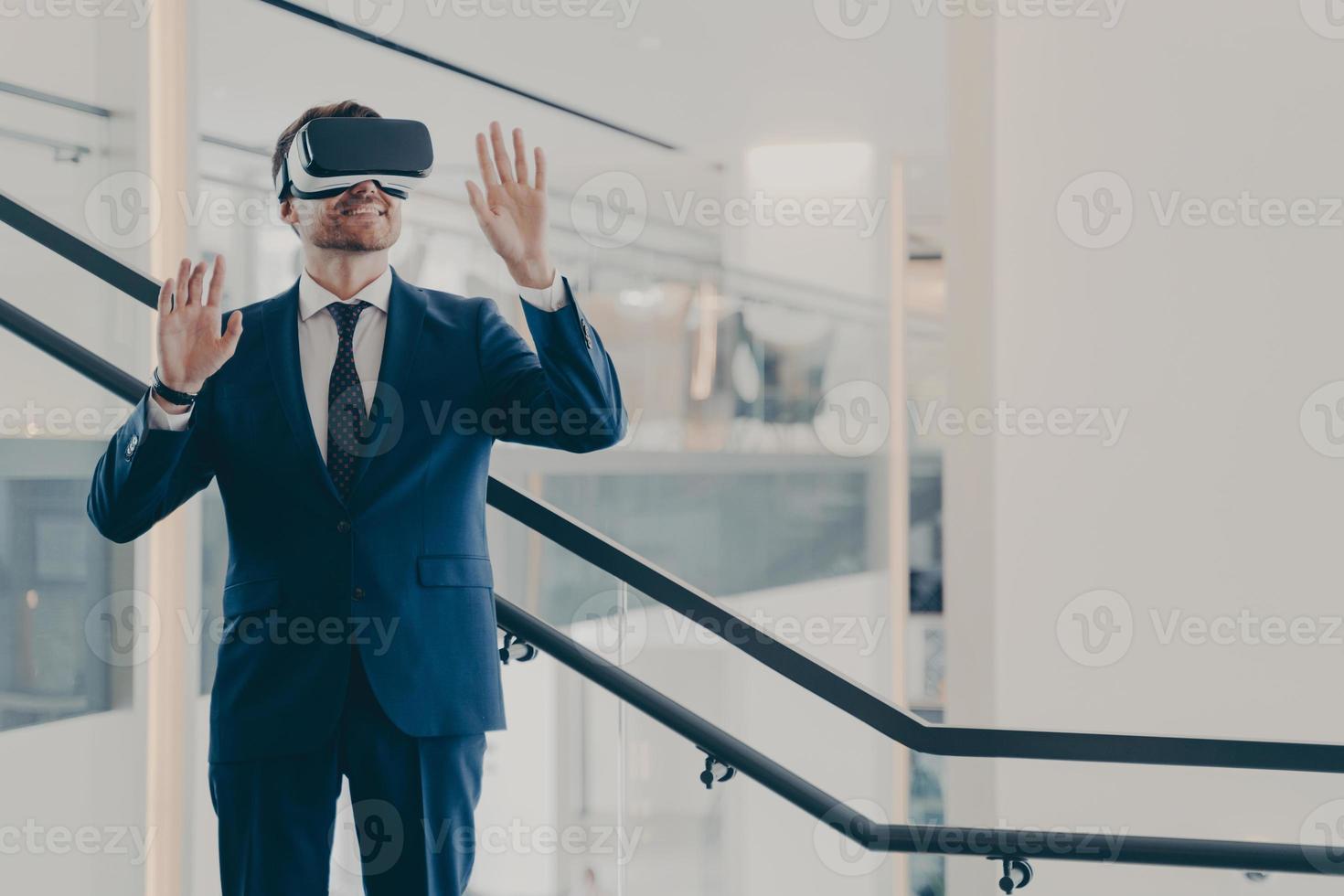 Impressed male office worker wearing VR goggles exploring virtual world while standing in office photo