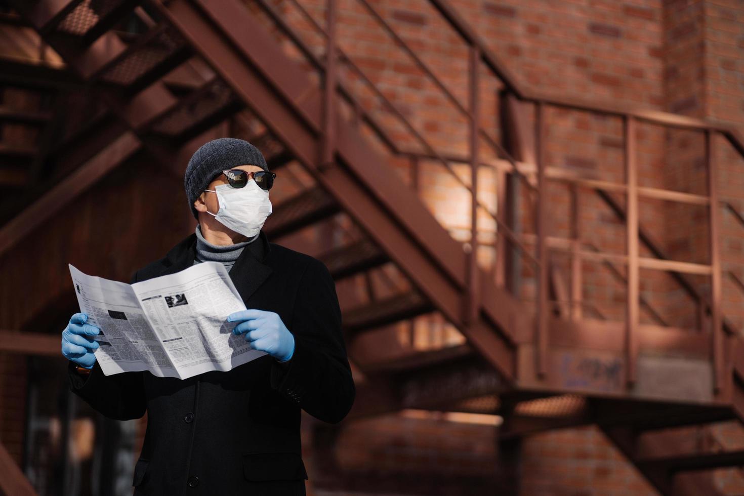 Horiontal shot of pensive man poses with newspaper, wears protective mask and rubber gloves, looks aside, thinks about self isolation, quarantine and coronavirus pandemic. People and covid 19 photo