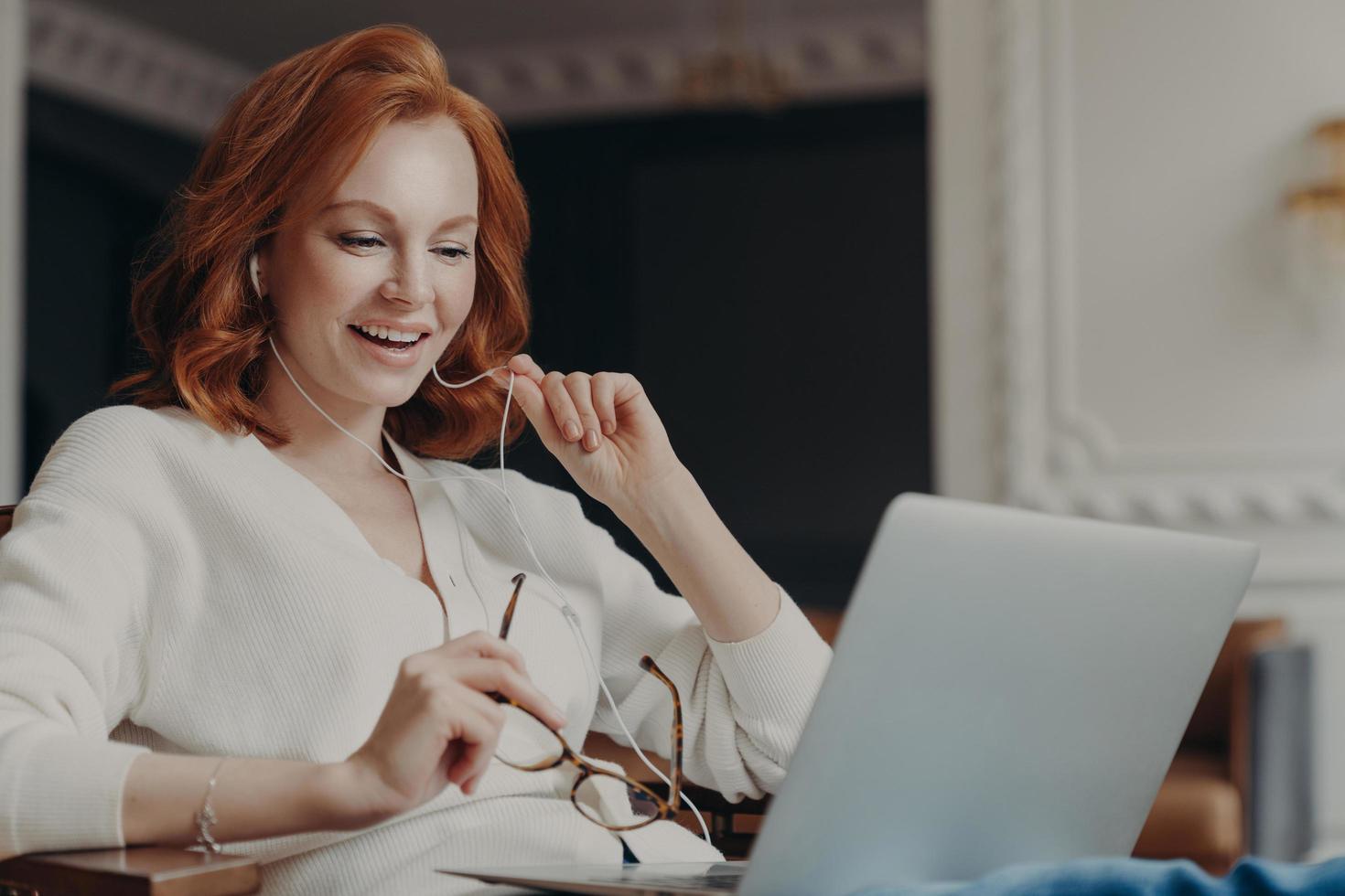 Satisfied happy foxy successful woman has video conference, uses modern laptop computer and earphones, browses data and works on remote project, poses in modern apartment, watches online webinar photo