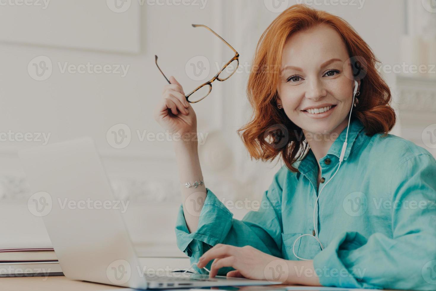 Glad red haired woman in blue shirt, works on laptop, uses earphones for watching webinar or online communication, listens courses, studies remotely from home during pandemic corona virus outbreak photo