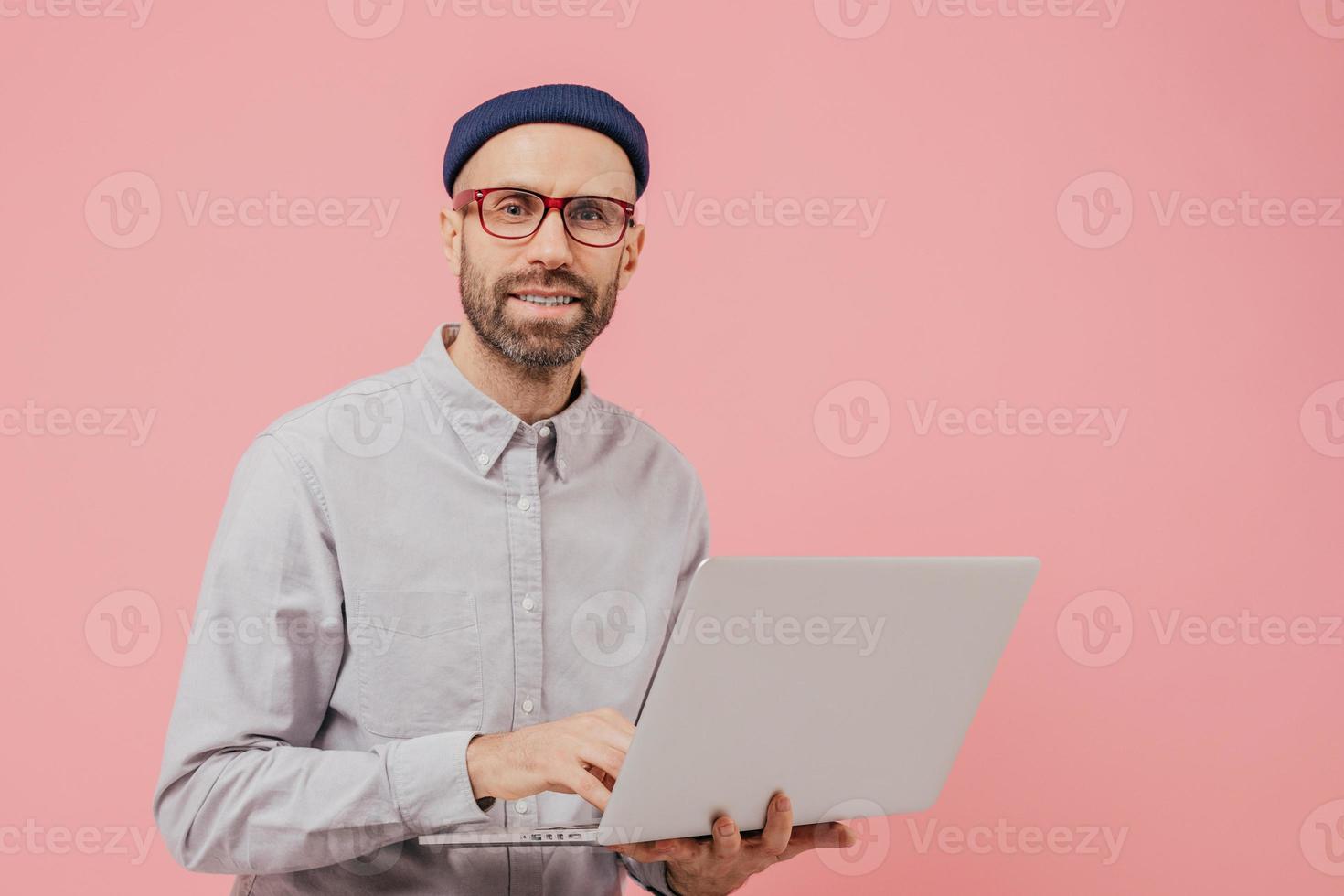 Photo of guy internet user browses website on laptop computer, connected to wireless internet, holds electronic gadget, wears spectacles, formal shirt, isolated over pink background with free space