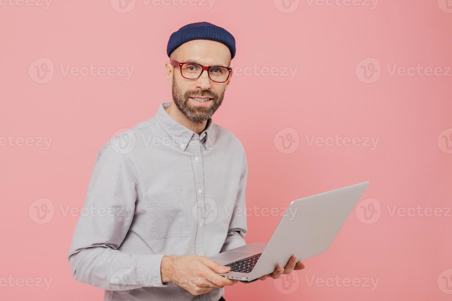 Talented male journalist with appealing look, holds modern laptop computer, works on new article, dressed in fashionable clothes, models against pink background, connected to wireless internet photo
