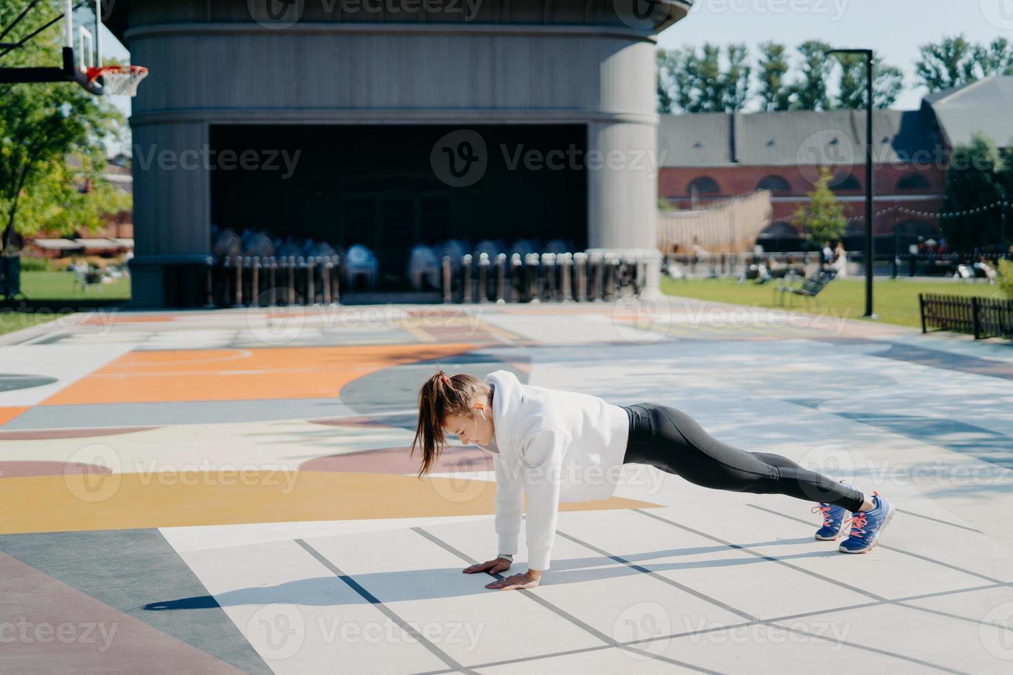 Full length side view shot of young active woman stands in plank position dressed in sweatshirt leggings sneakers has training outdoors on stadium goes in for sport. Morning workout concept. photo