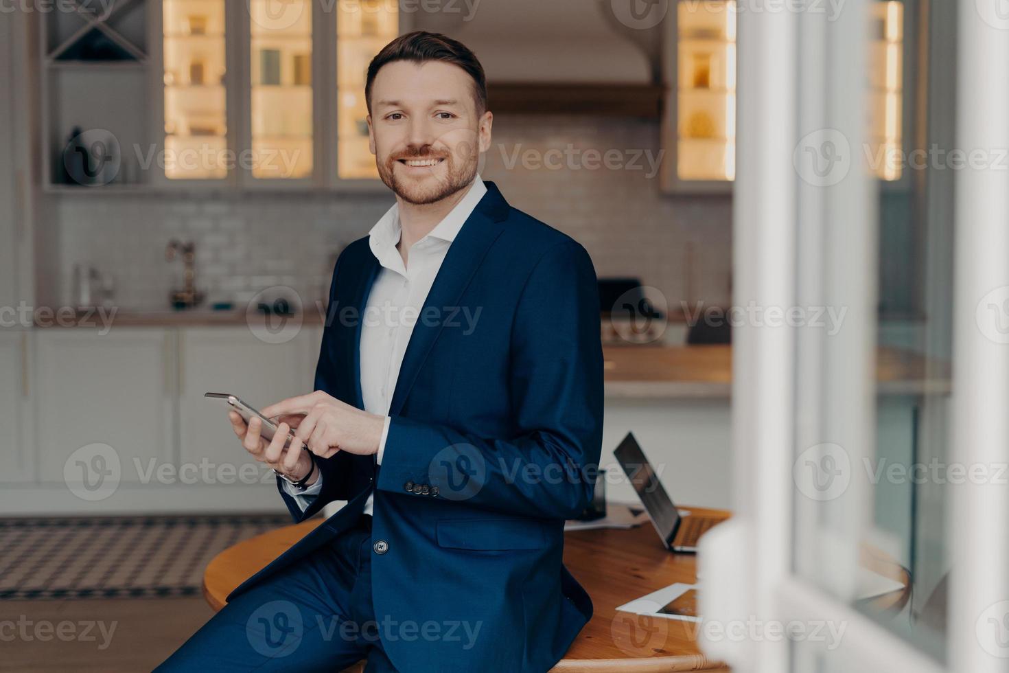 Horizontal shot of man employee uses great business app reads good news message holds mobile phone poses against kitchen interior near desktop wears formal outfit smiles happily. Mobile payments photo
