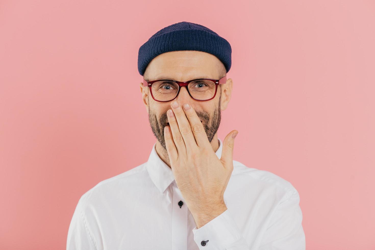 Positive unshaven man covers mouth with palm, giggles positively, wears spectacles, hears funny joke, models against pink background. Happy male designer recieves good news. Emotions concept photo