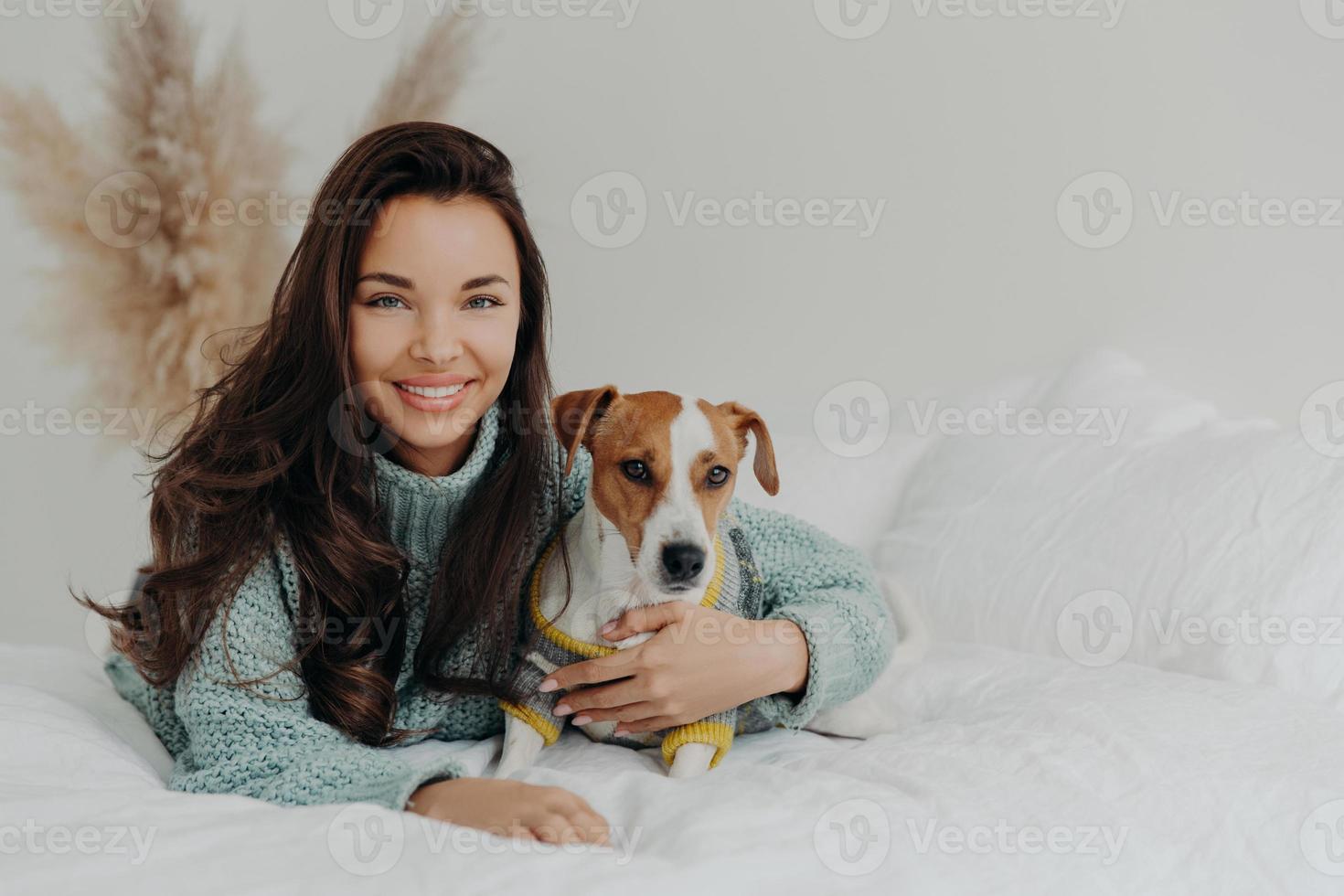 Photo of cheerful woman embraces dog with love, spend free time together, expresses tender feeling and emotions, falls in love with pet, lie on comfortable bed. Positive emotions, animals and care