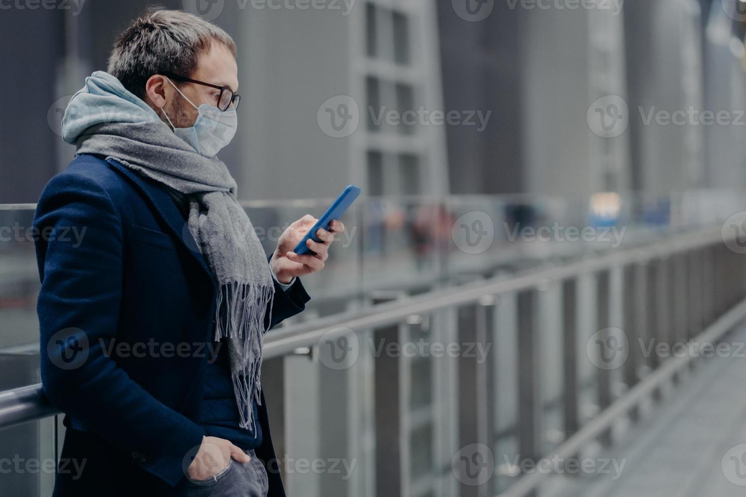 Serious European man looks in smartphone, reads news online, sends text messages, being protected from virus, wears medical mask, cares about public coronavirus protection. Virus outbreak, Covid-19 photo