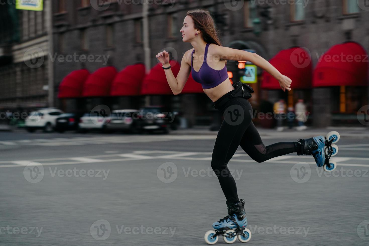 Horizontal shot of active slim young woman rides fast on rollerskates improves balance agility and coordination dressed in cropped top and leggings poses on street road focused into distance photo