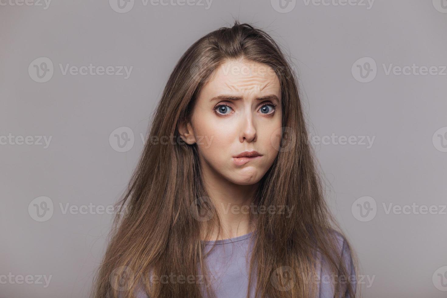Offended stressful female bites lower lip with dissatisfaction, has sorrorful expression, expresses negative emotion, being in low spirit after quarrel with somebody, isolated over grey background photo