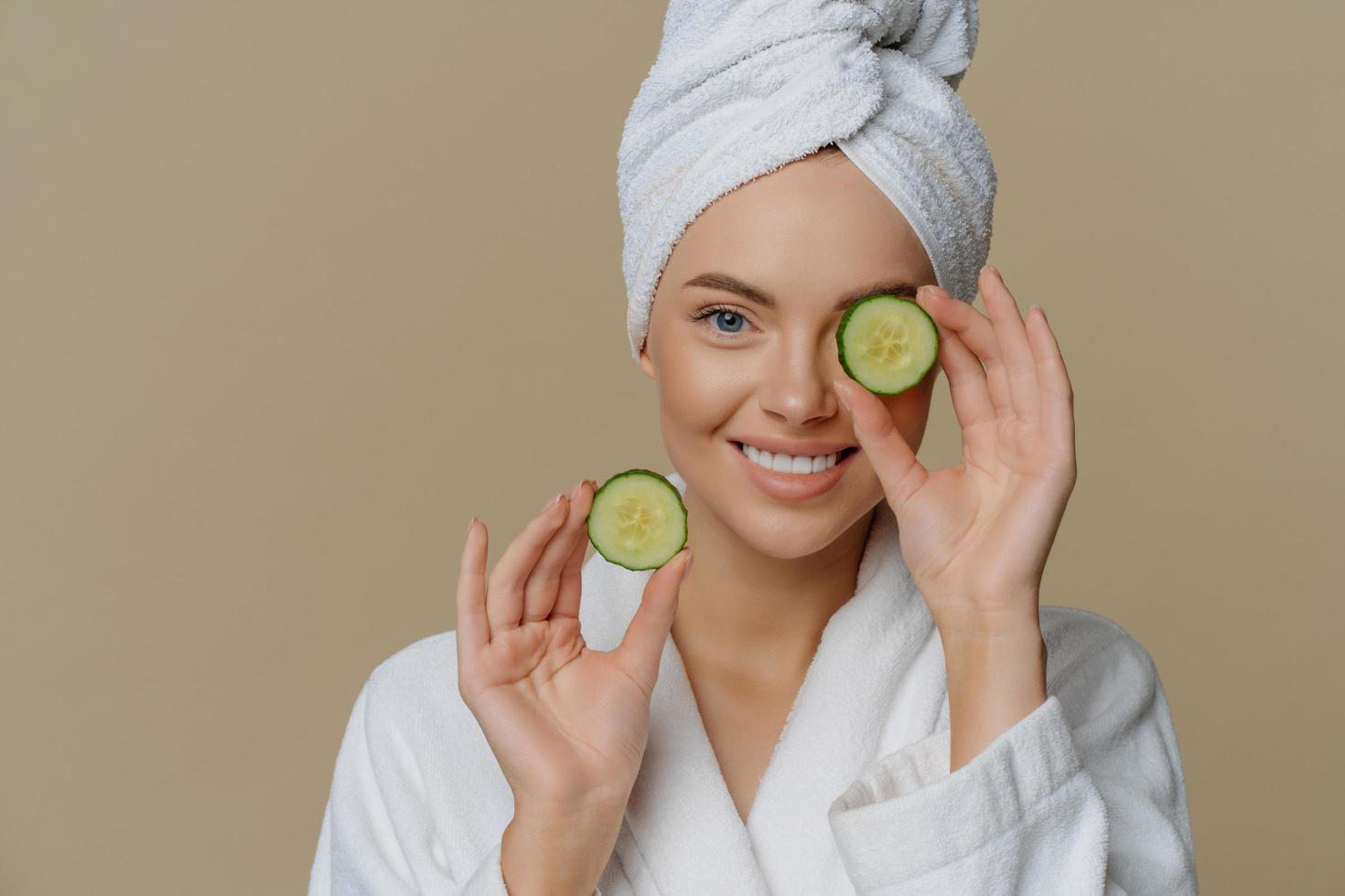 Skin care natural beauty and cosmetology concept. Positive refreshed beautiful woman holds slices of fresh cucumber makes mask for face skin dressed in domestic robe wrapped bath towel on head photo