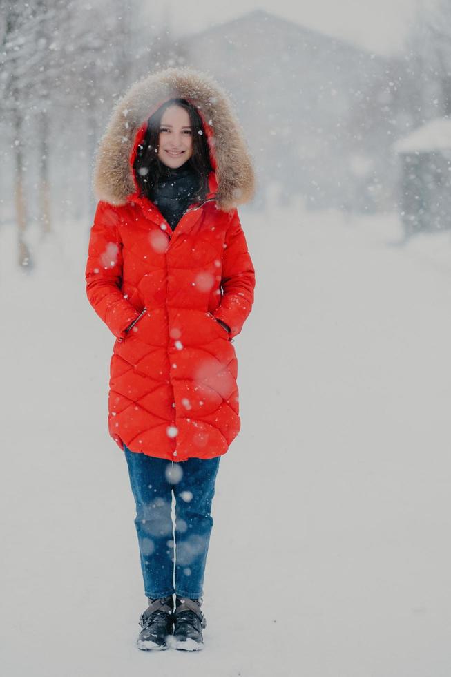 Full length vertical shot of pretty woman wears red winter jacket, jeans and boots, keeps hands in pockets, stands on street during winter time during snowflake, poses for making photo outdoor