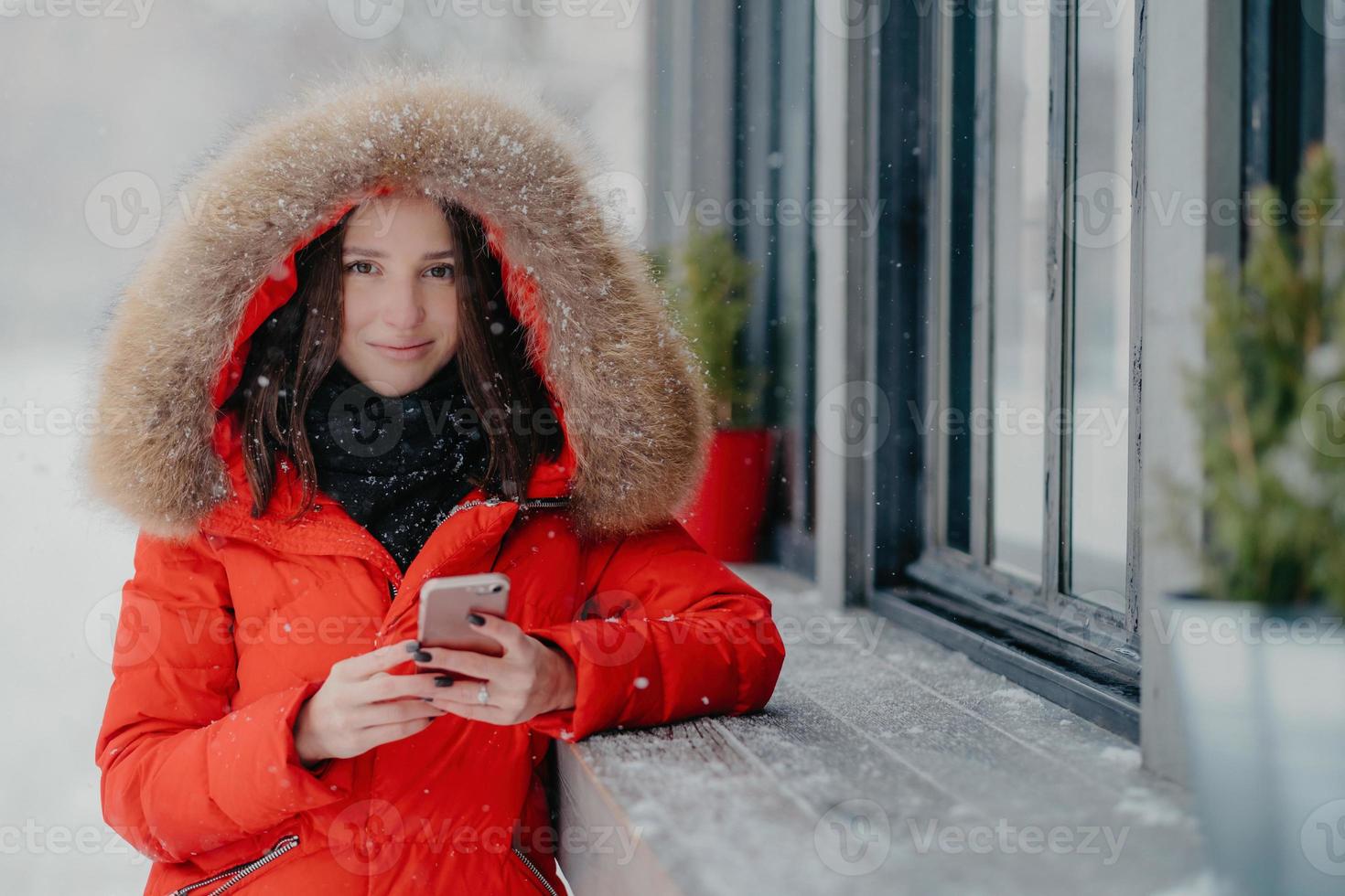 Smiling young European female in red coat, has black manicure, uses modern electronic gadget for communication in internet with friends, likes winter weather, sends photo in social networks.