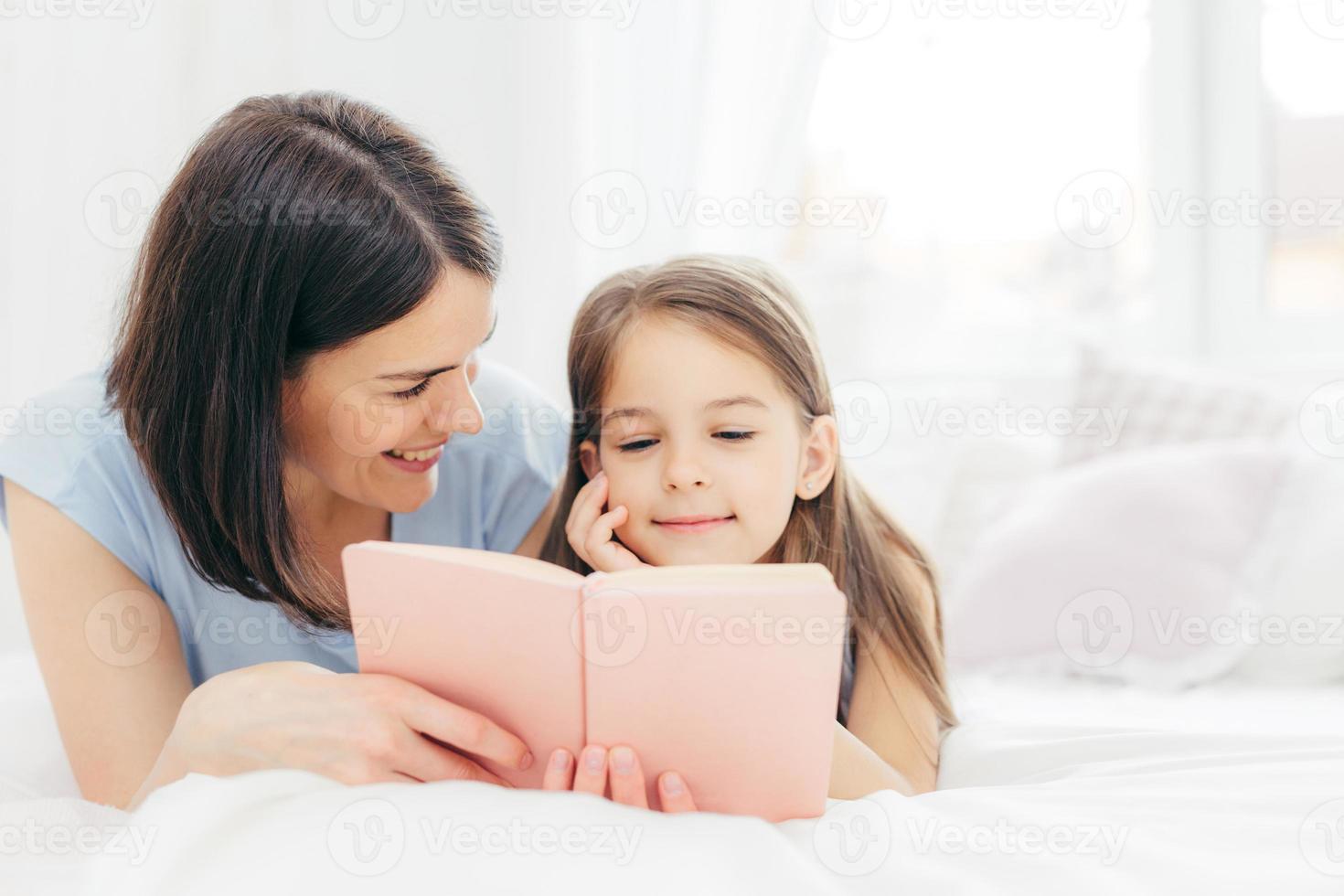 afectuosa madre joven con cabello oscuro, mira felizmente a su pequeña hija, lee un libro interesante o un cuento de hadas juntos, disfruta del ambiente doméstico en casa. gente, familia, concepto de infancia foto