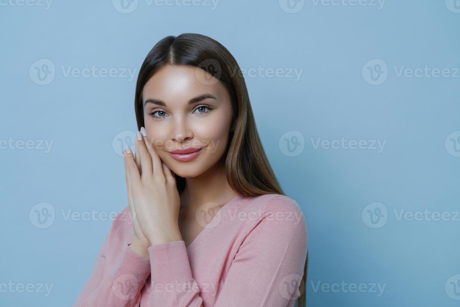 Close up portrait of pleased attractive brunette woman gazes directly at camera, keeps hands near face, listens advice from cosmetologist how to care about skin, models indoor against blue wall photo