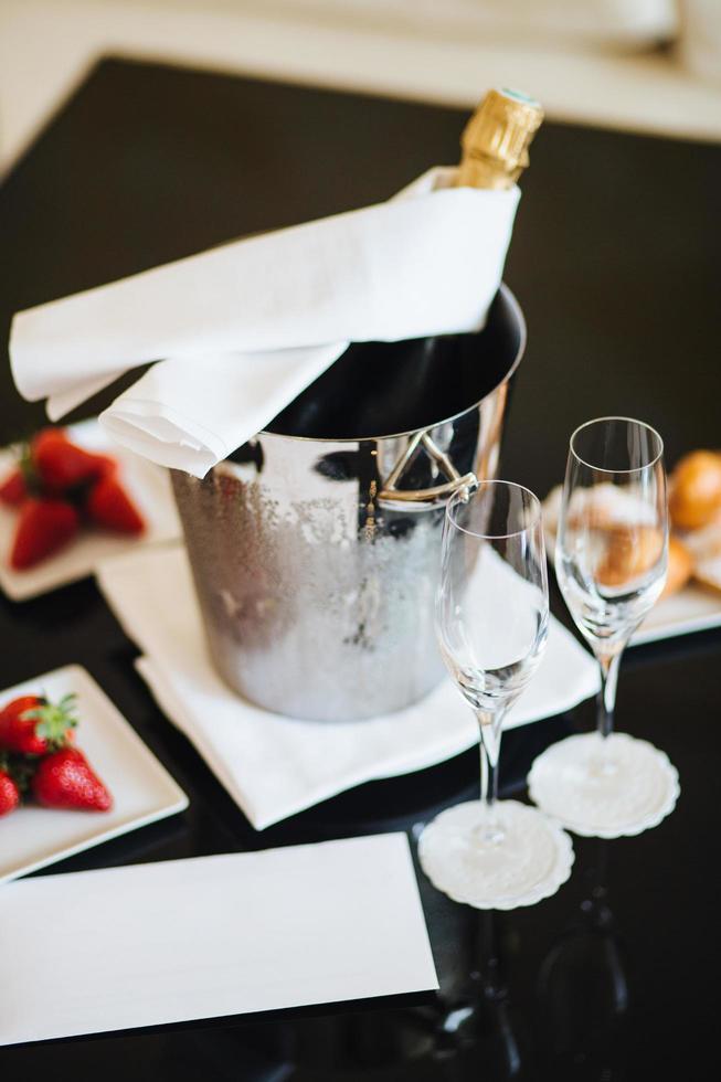 Vertical shot of bottle with alcoholic drink in busket to be cooler, two glasses and dessert for celebration something. Tasty juicy strawberry and champagne. Celebration. photo