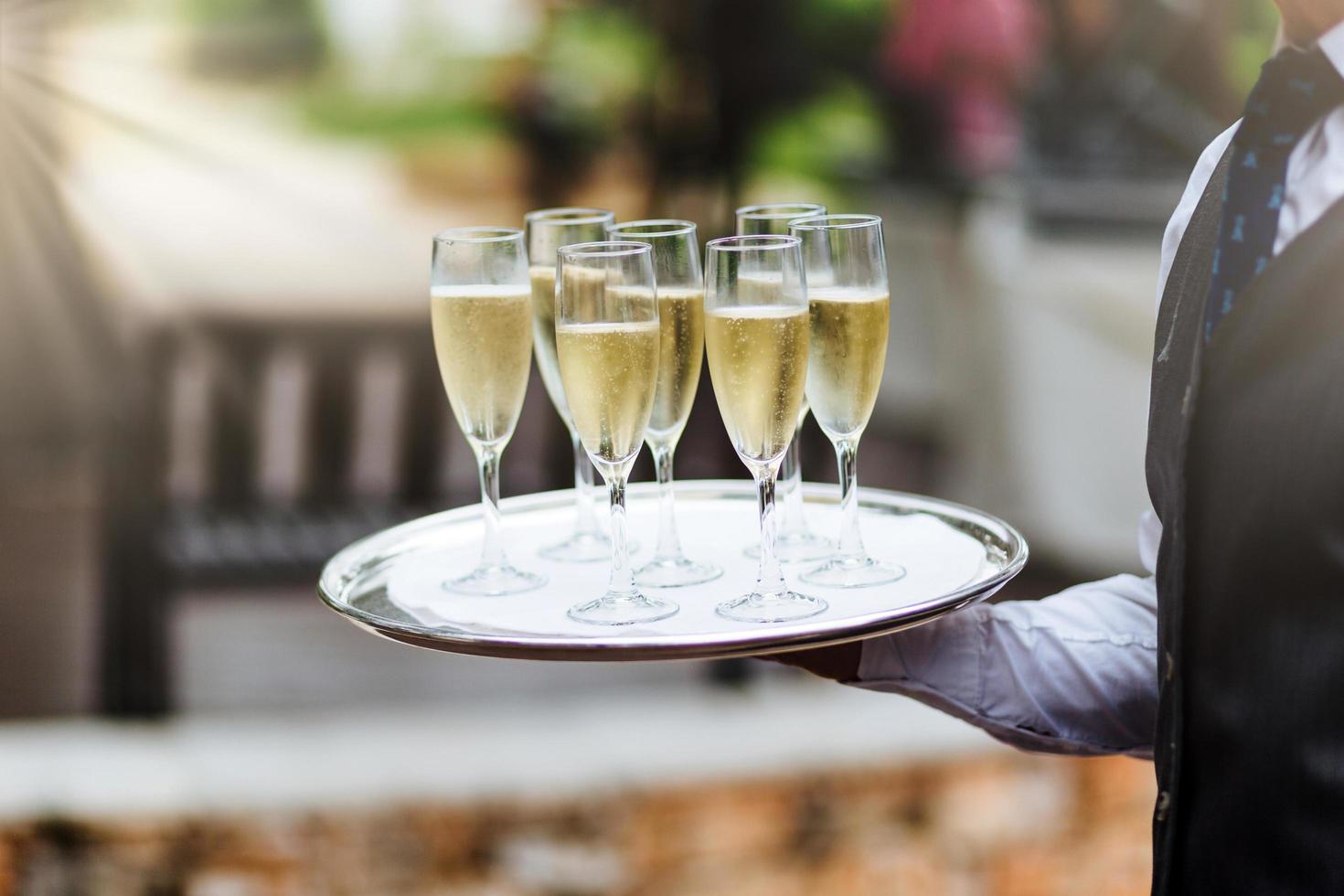 Unrecognizable waiter in formal clothing holds tray with glasses of champagne, carries drinks on festive table to guests, supplies good service for visitors. Hospitality, service and drinking concept photo