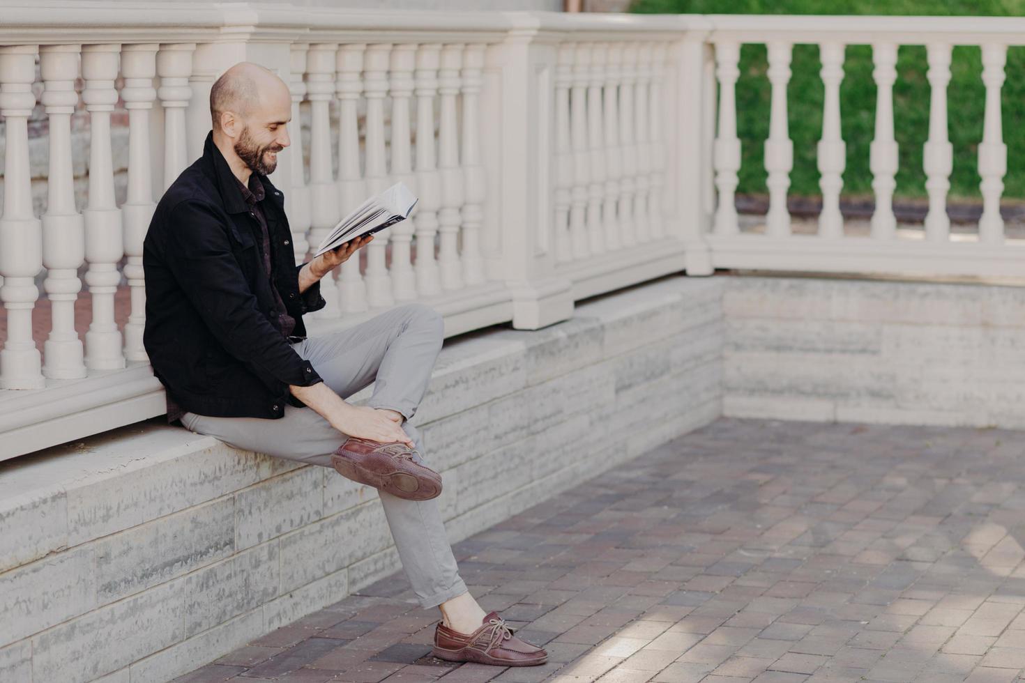 retrato al aire libre de un hombre feliz de mediana edad con una apariencia atractiva, sostiene un libro, lee algo divertido, disfruta del tiempo libre en el parque, posa contra el interior del balcón blanco. concepto de personas y pasatiempos foto