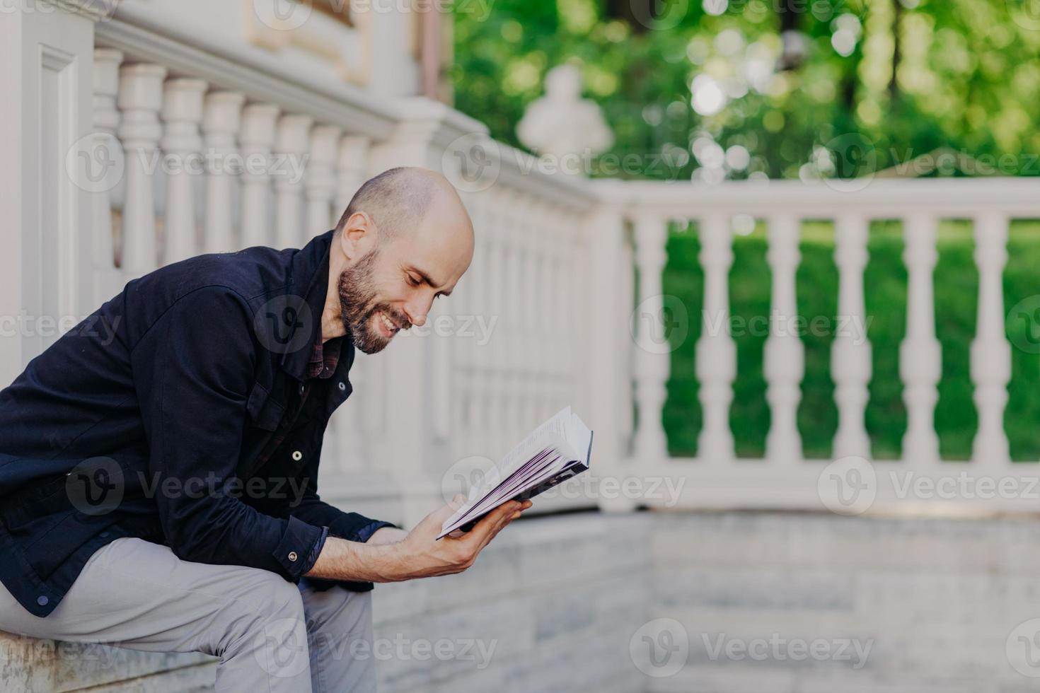 People, hobby and lifestyle concept. Postive bald man reads book with enthusiasm, poses outdoor against white balcony, has happy expression, enjoys calm atmosphere. Middle aged handsome male studies photo