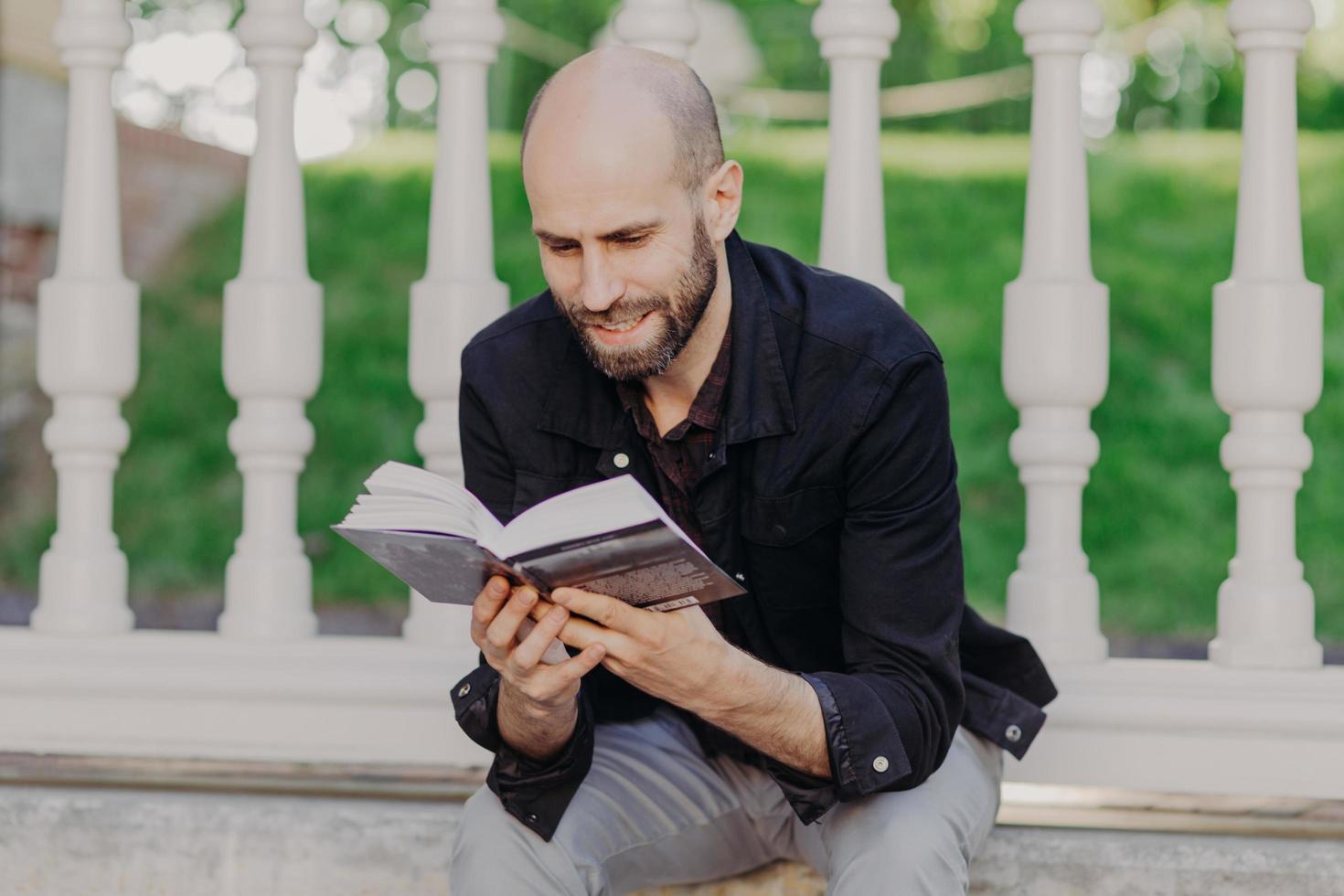 Close up shot of focused attractive bald young man holds book, reads with joy, enjoys weekends, spends free time in summer garden, likes reading in calm atmosphere. People and relax concept. photo