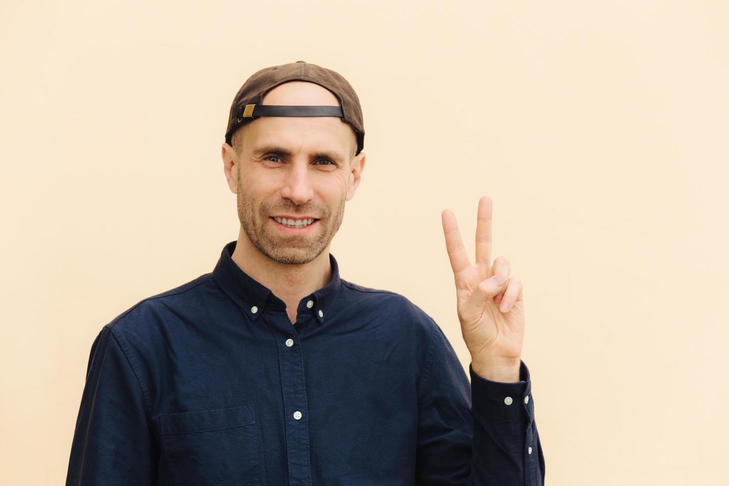 Portrait of handsome delighted unshaven male shows peace sign, keeps two fingers raised, dressed in cap and shirt, isolated over light background. Cheerful successful young man gestures indoor photo