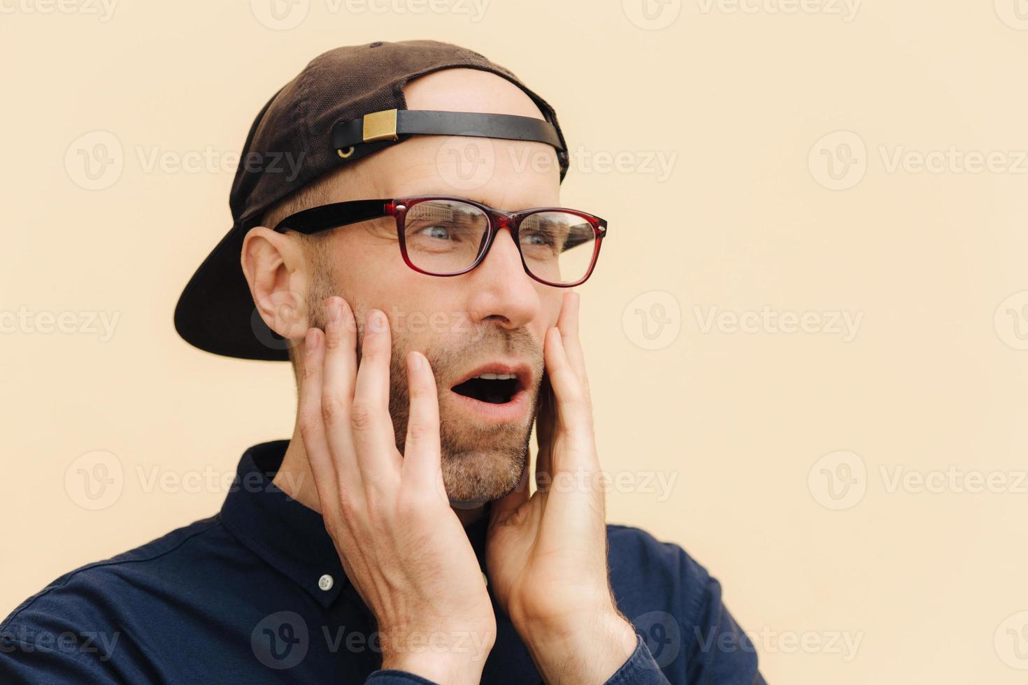 Close up portrait of attractive male with stubble, wonders something and looks with surprisement aside, keeps hands on cheeks, wears glasses and fashionable cap, isolated over light background photo