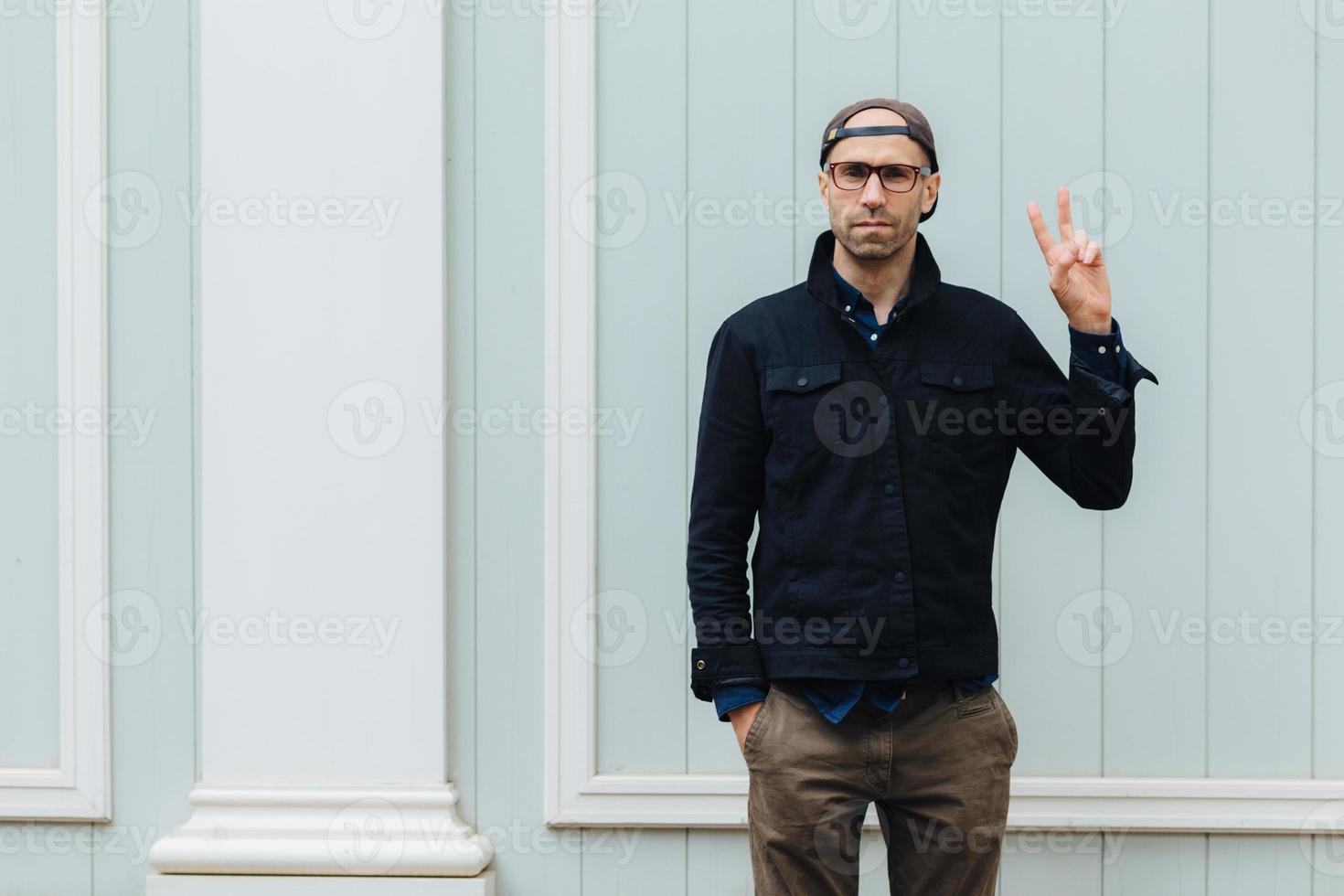 Portrait of handsome of unshaven serious fashionable male shows peace sign, wears spectacles and stylish clothes, keeps hand in pocket, poses against white wall outdoor. Body language concept photo