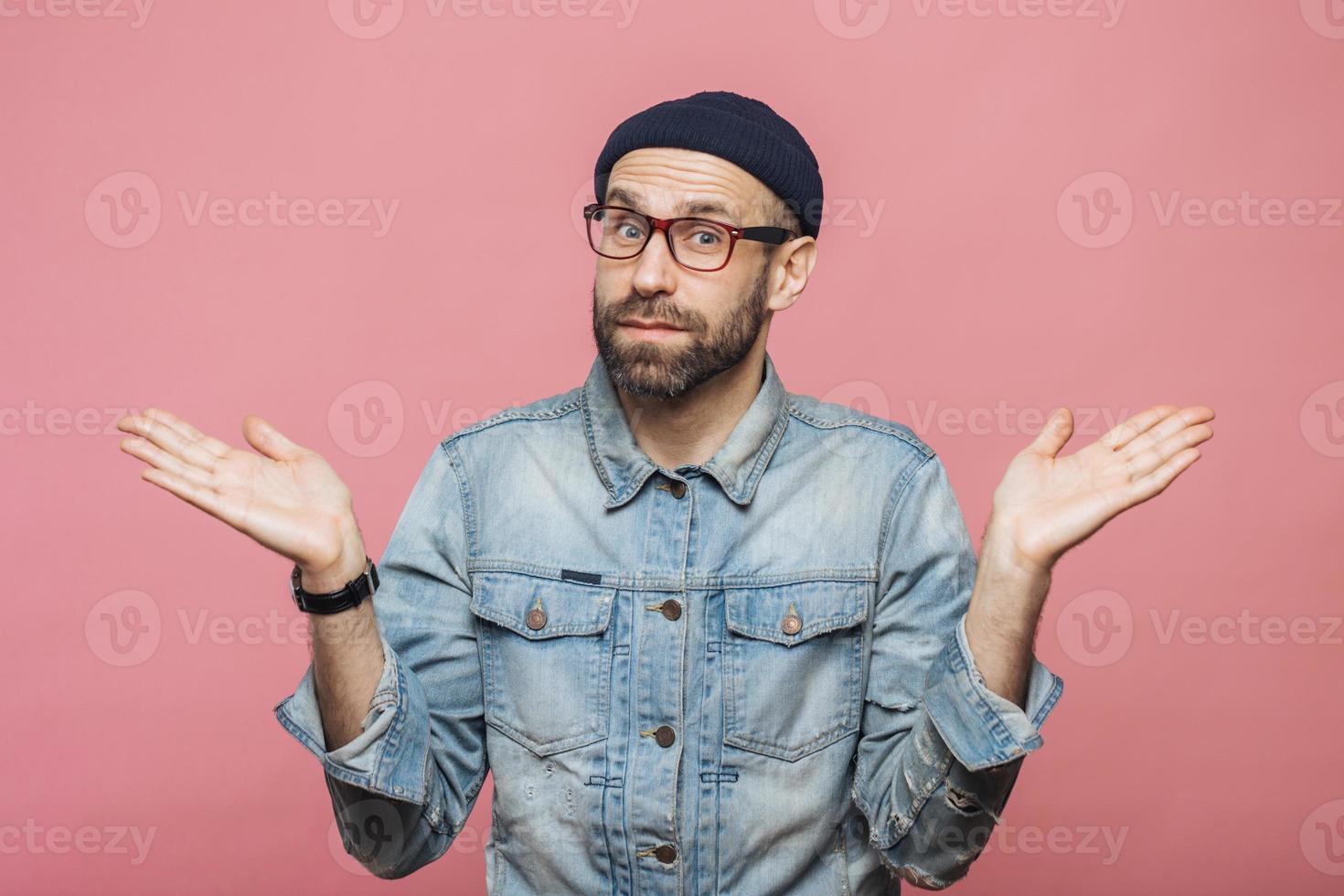 Indoor shot of hesitant bearded man shrugs shoulders, looks uncertain, doubts when makes decision, isolated over pink background. People, doubt and hesitation concept. Body language concept. photo