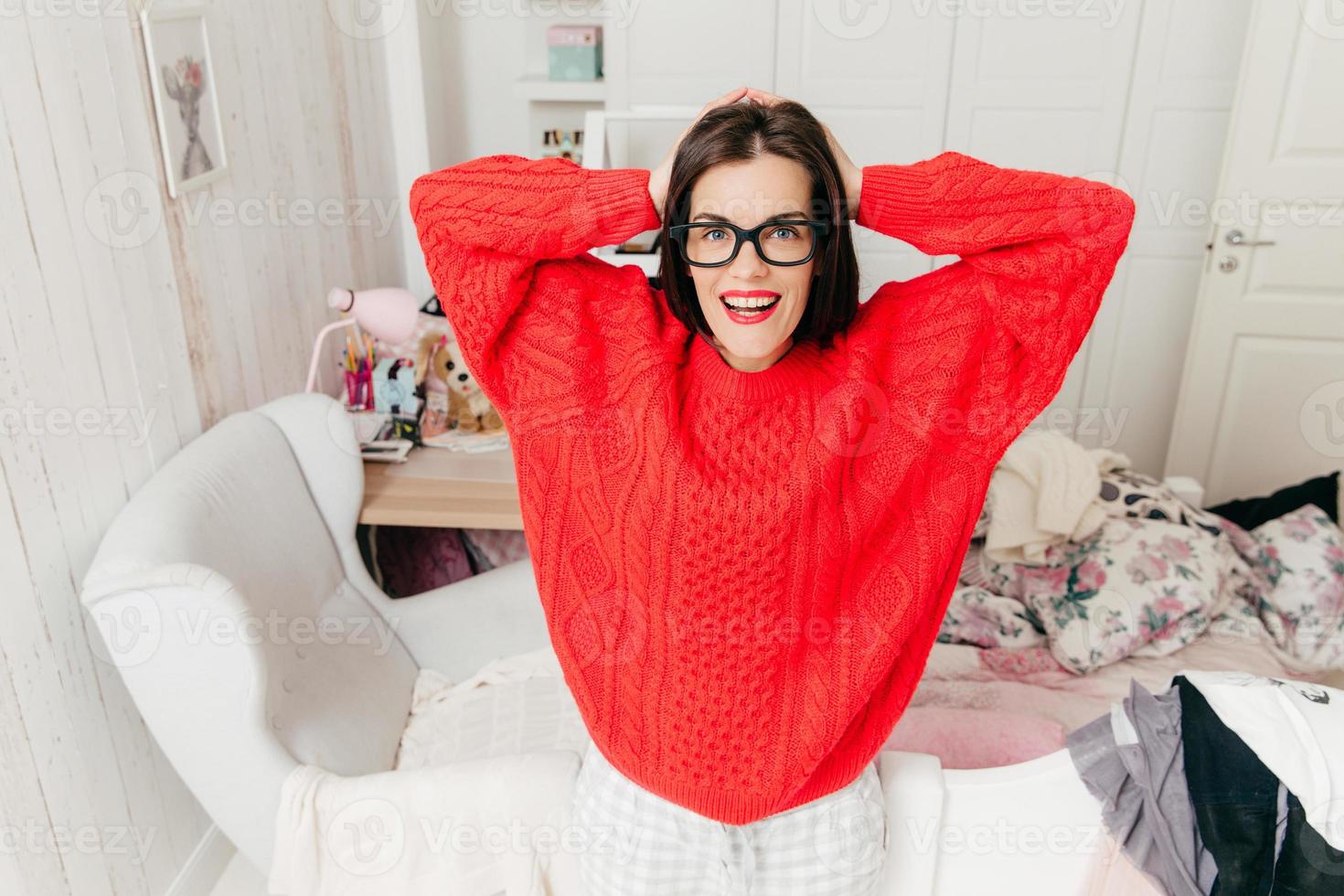 Horizontal portrait of cheerful brunette housewife wears red warm sweater and spectacles, poses against home interior in her room, has positive smile. Beautiful woman with happy expression indoor photo