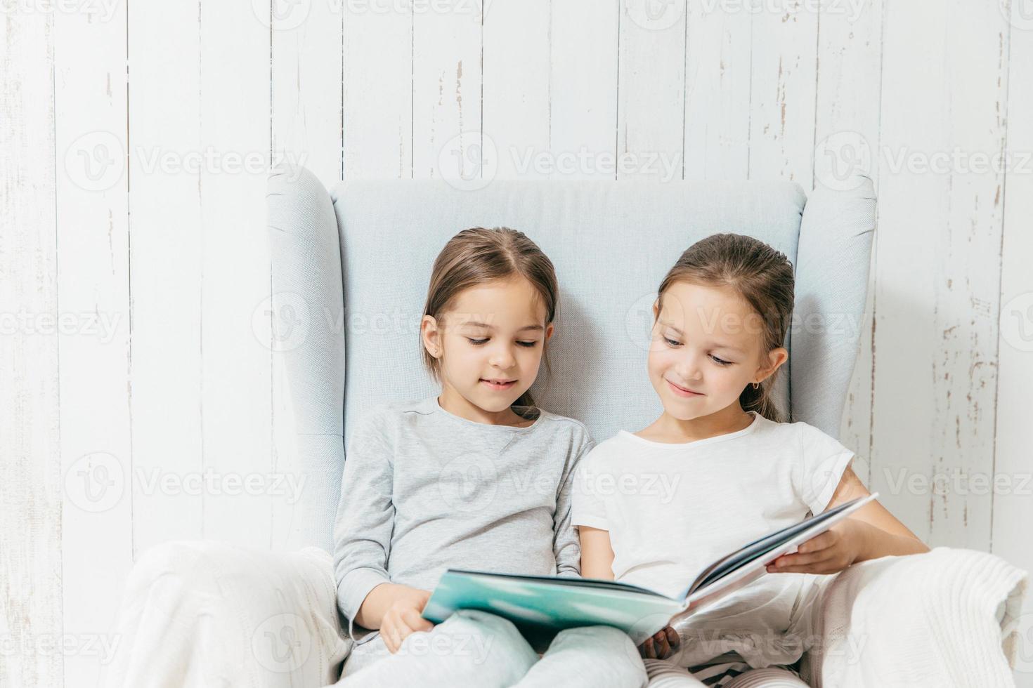 Two little adorable sisters sit in sofa, read interesting book, sit in comfortable sofa, enjoy togetherness. Best friends female children hold book, view pictures and read hearders, have spare time photo