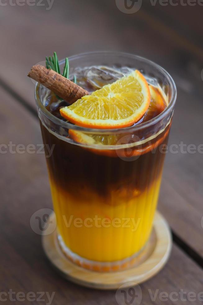 café negro helado de primer plano mezclado con jugo de naranja en un vaso con naranjas en rodajas, hojas de romero y canela en la parte superior. en un posavasos de madera colocado sobre una mesa de madera. foto