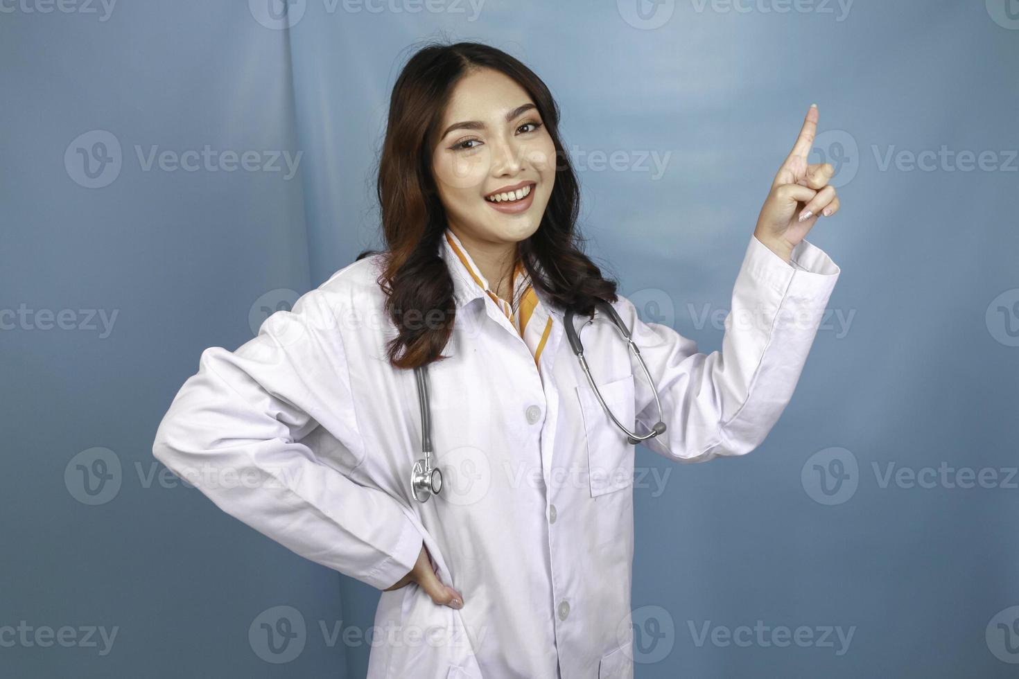 Portrait of a young Asian woman doctor, a medical professional is smiling and pointing upward at a copy space isolated over blue background photo