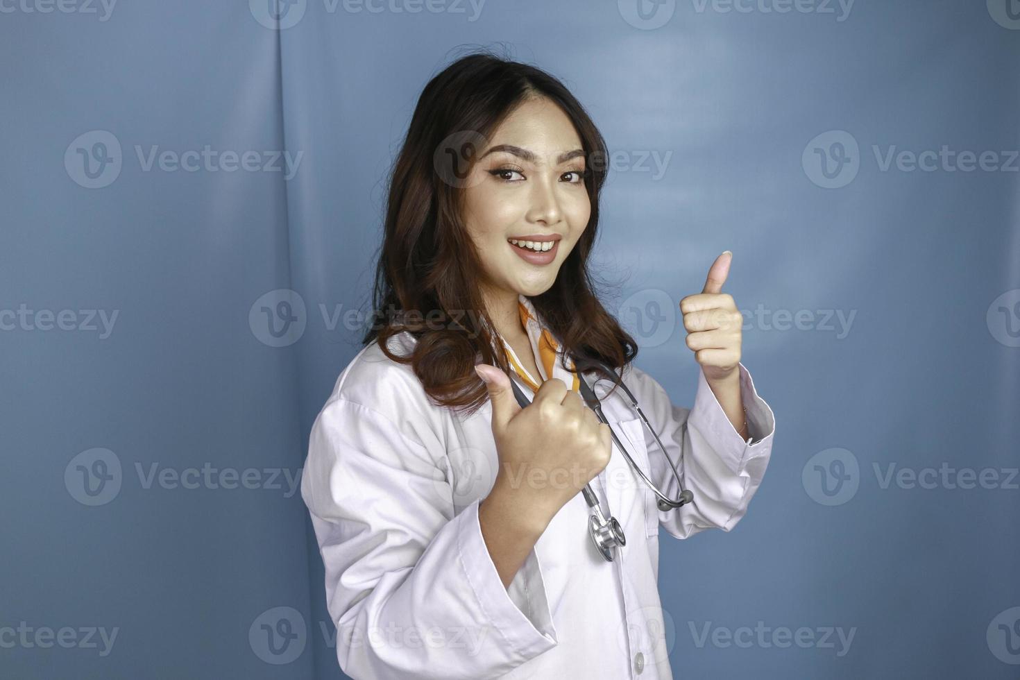 retrato de una joven doctora asiática, un profesional médico sonríe y muestra el pulgar hacia arriba o el signo de ok aislado sobre fondo azul foto