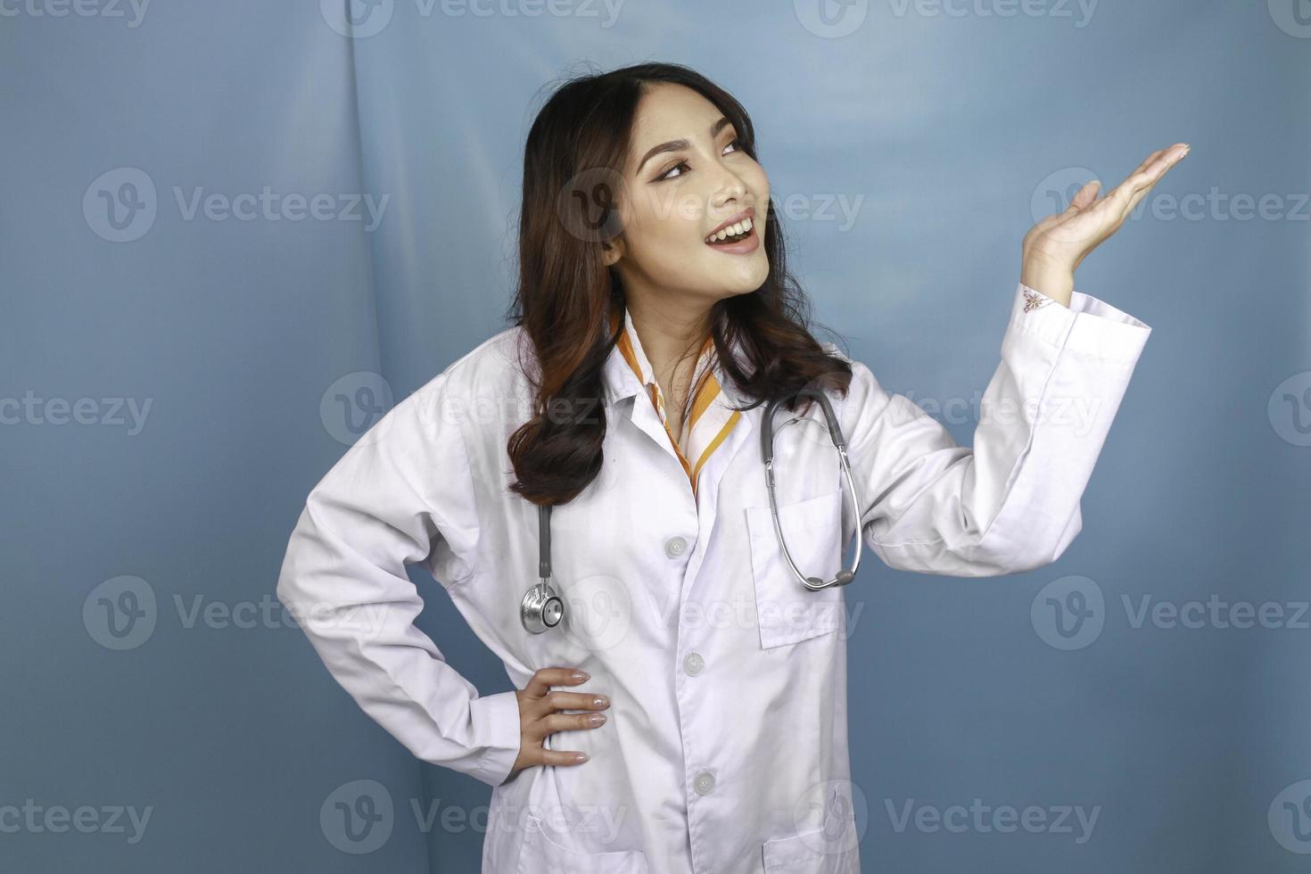 Portrait of a young Asian woman doctor, a medical professional is smiling and pointing upward at a copy space isolated over blue background photo