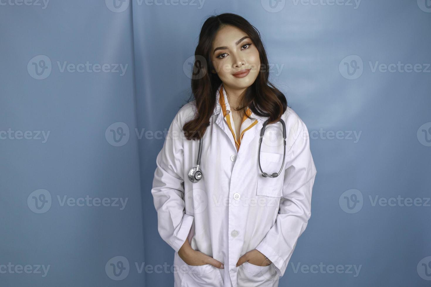 Confident relaxed female Asian doctor standing with hands in the pockets of her lab coat smiling quietly at the camera over blue background with copy space photo